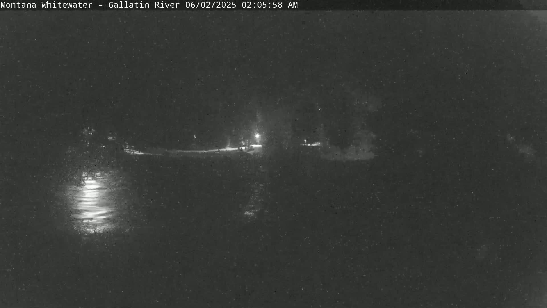 A nighttime, low-light view of a dark body of water reflecting light, with a dark shoreline and dimly lit area beyond.