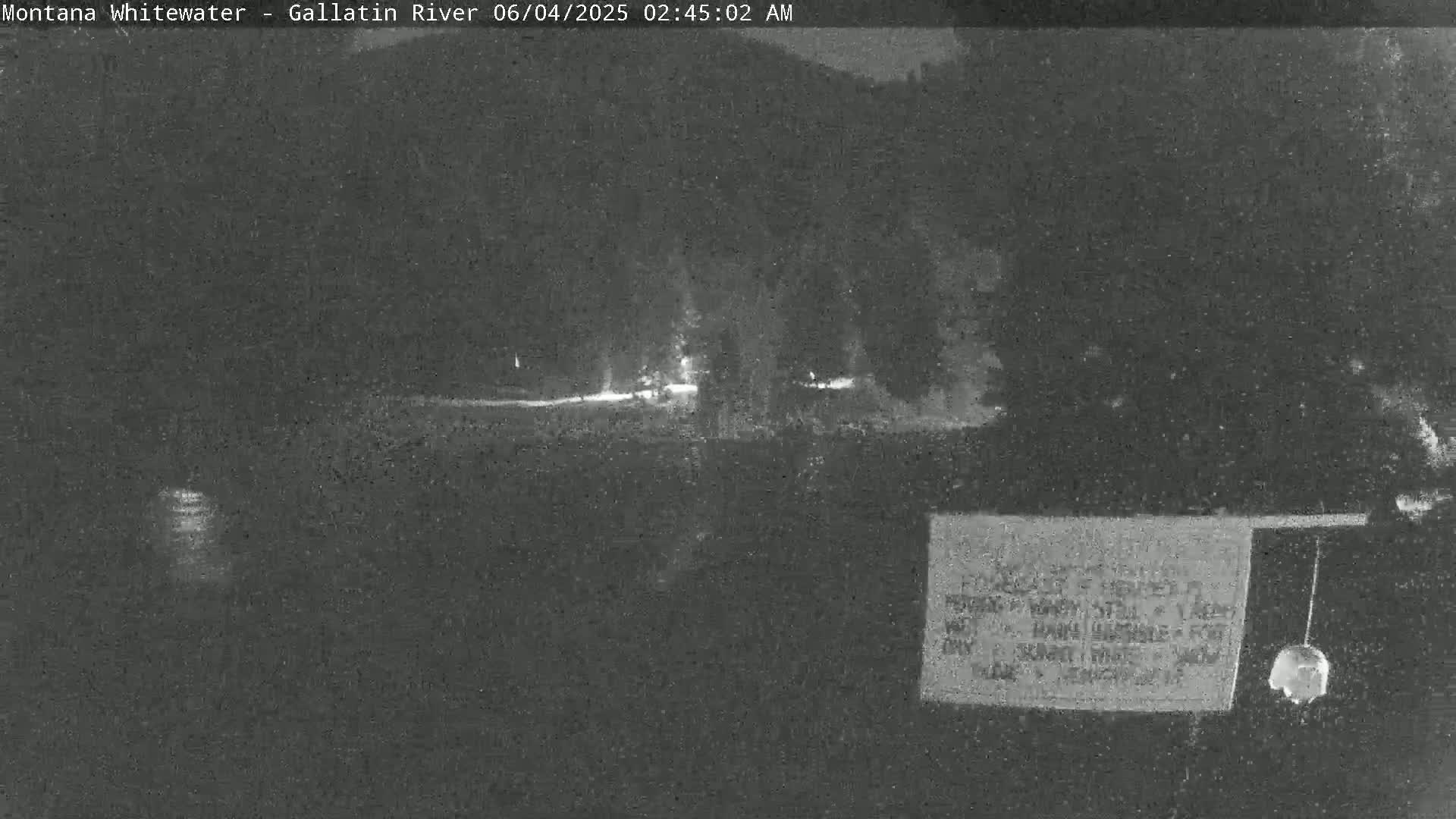 A nighttime, low-light image shows a dark, wooded area with a faintly visible road or path and some distant lights.
