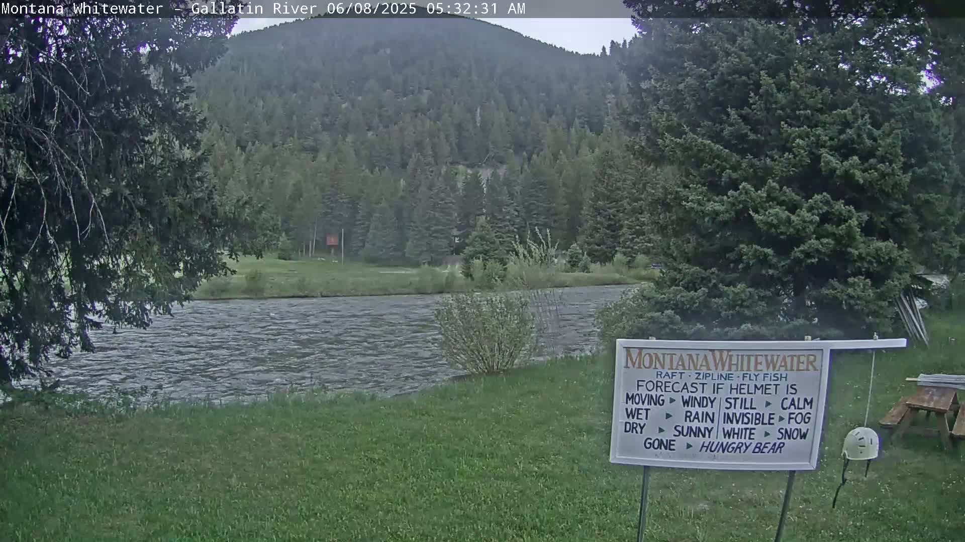 A calm river flows past a grassy bank beneath a cloudy sky, with a forested hillside rising in the background.