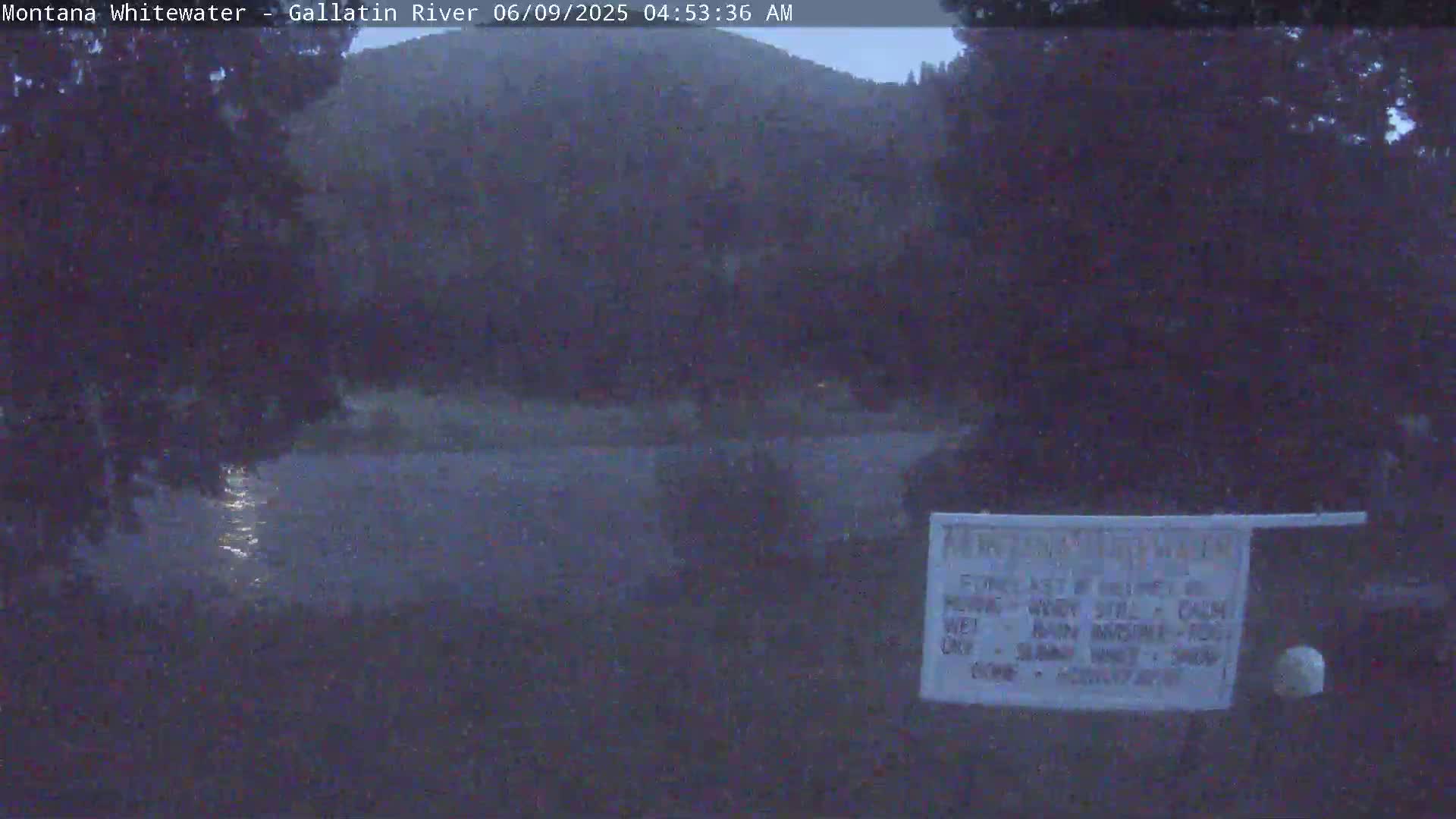 A dark, calm river reflects light at its center, with trees and a hill visible in the background under a predawn sky.