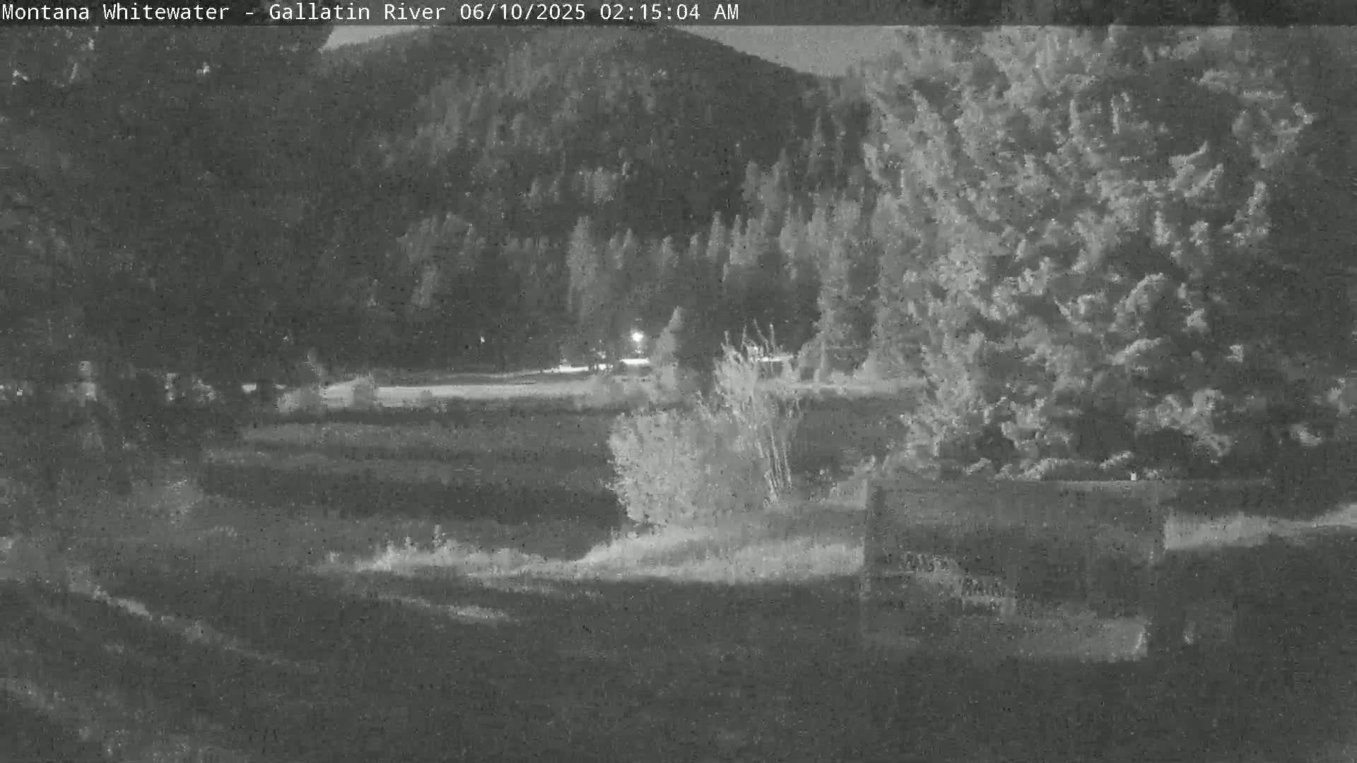 A nighttime, low-light view of a grassy field and trees near a road, with a dark hillside in the background.