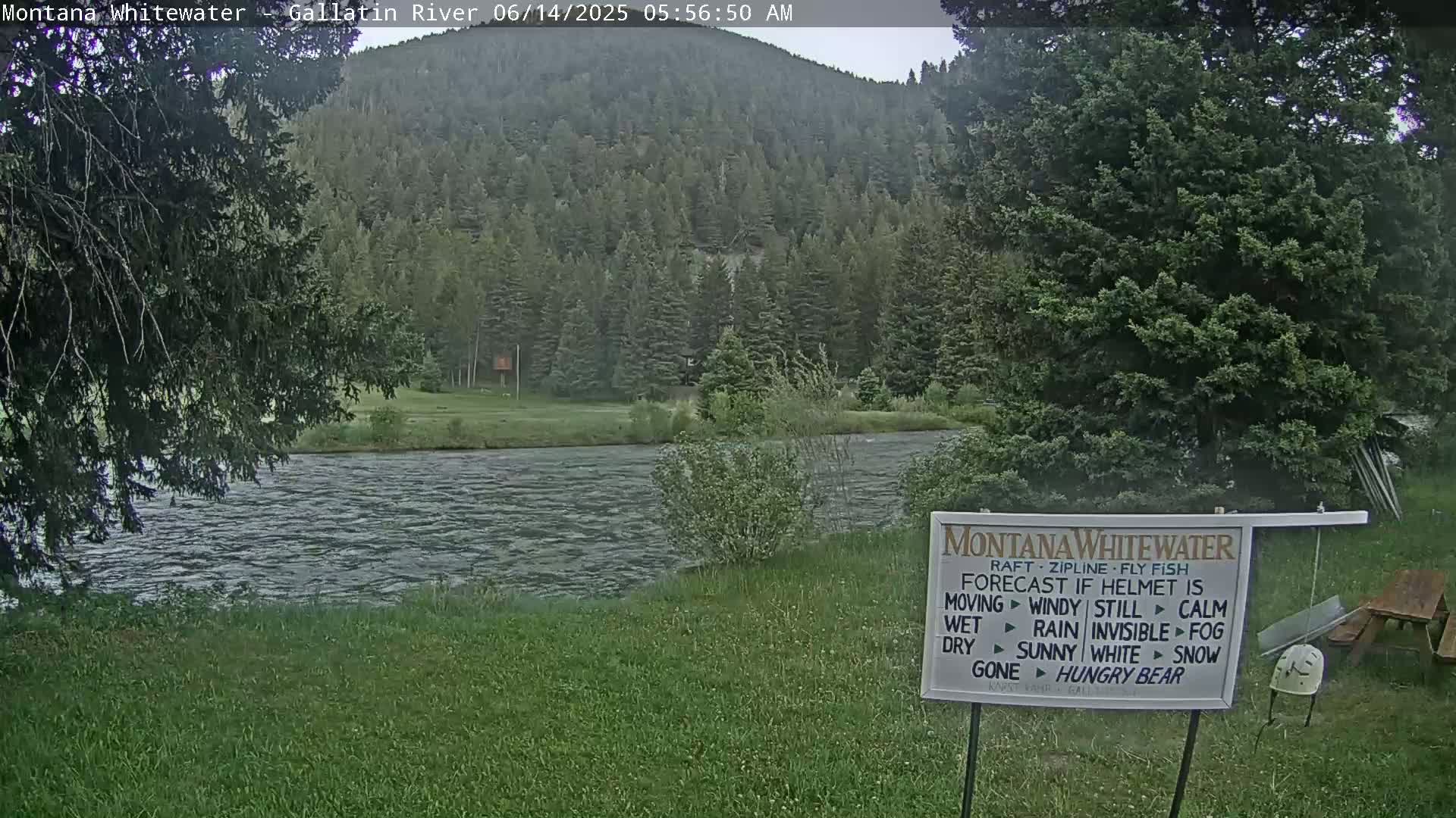 A calm river flows beneath a verdant, forested hill under an overcast sky.