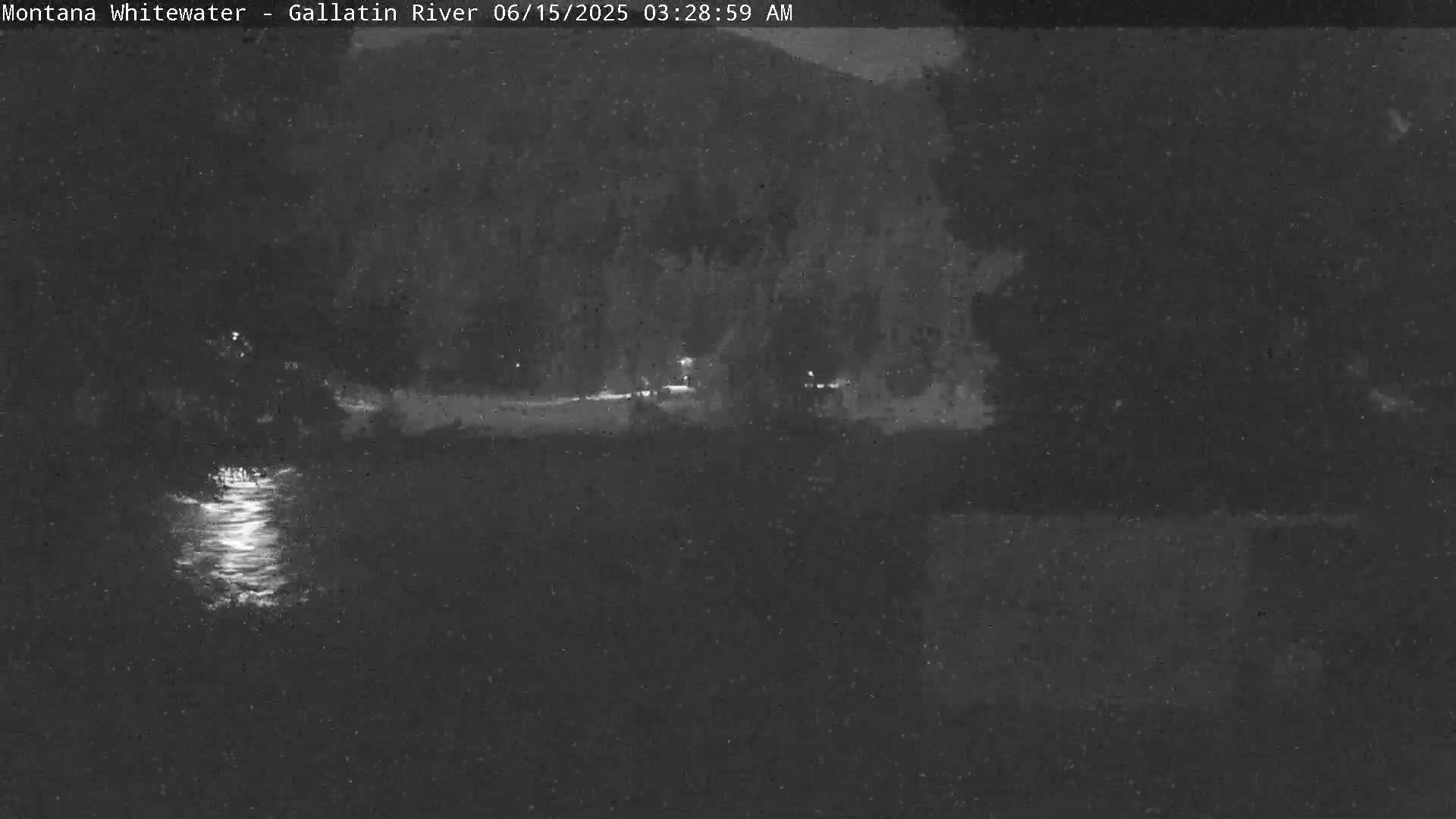 A nighttime, low-light view of a river reflecting light, with dark trees and land in the background.