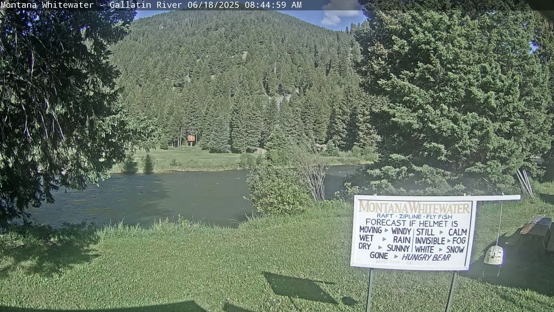 A calm river flows under a partly sunny sky, past a grassy bank and a lush green forest.
