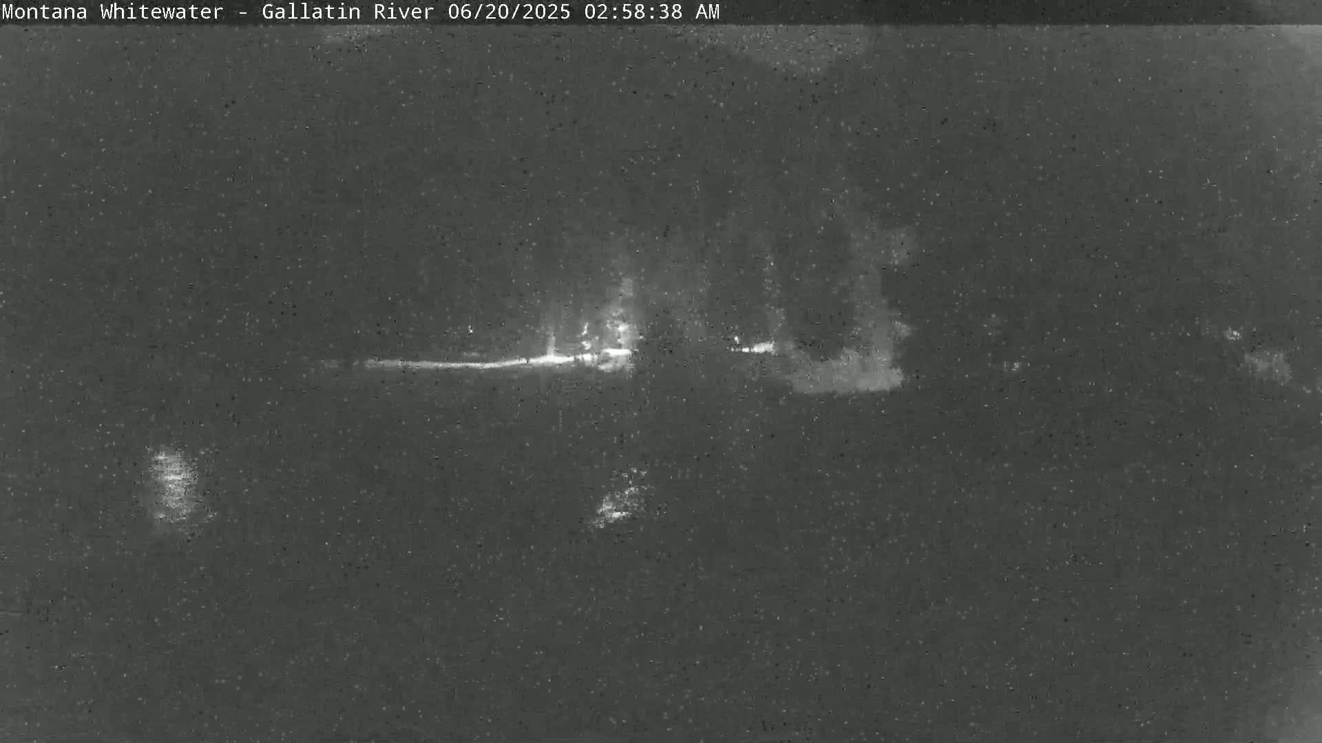 A nighttime, low-light image shows a dark, flat landscape with a faint, horizontal line suggesting a road or riverbank, and sparsely distributed bright spots of light in the distance.