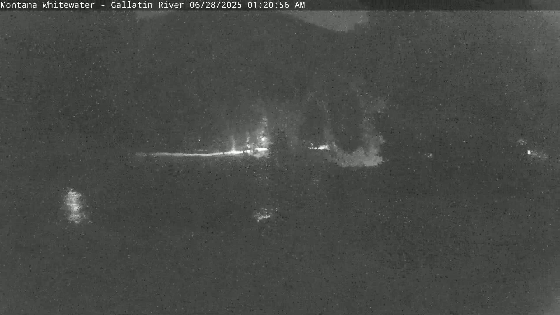 A nighttime, low-light image shows a dark landscape with a faintly lit area possibly indicating a road or riverbank.