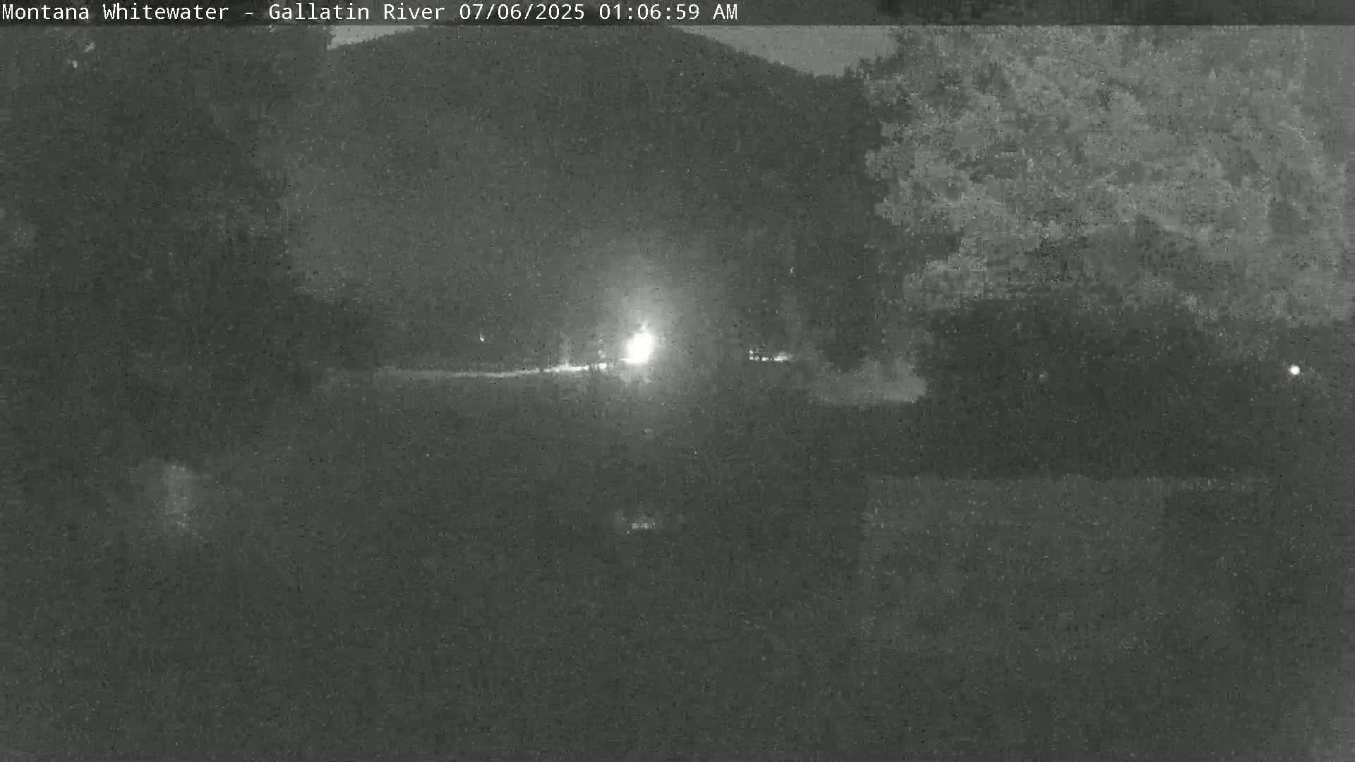 A nighttime, low-light view of a dark, possibly wooded area with a bright light source in the distance.