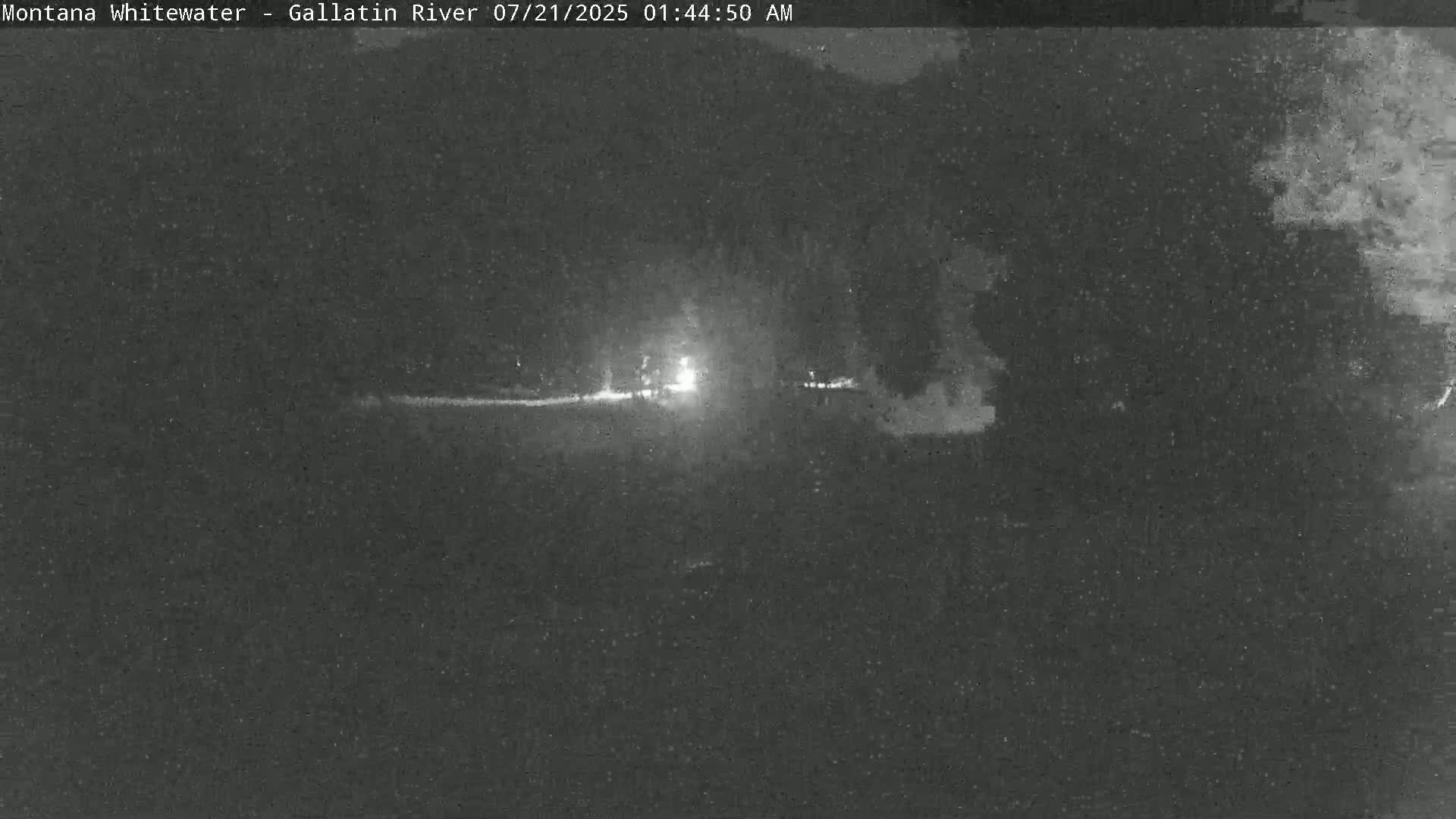 A nighttime, low-light view of a dark, possibly rural landscape with a faintly illuminated area in the middle distance.
