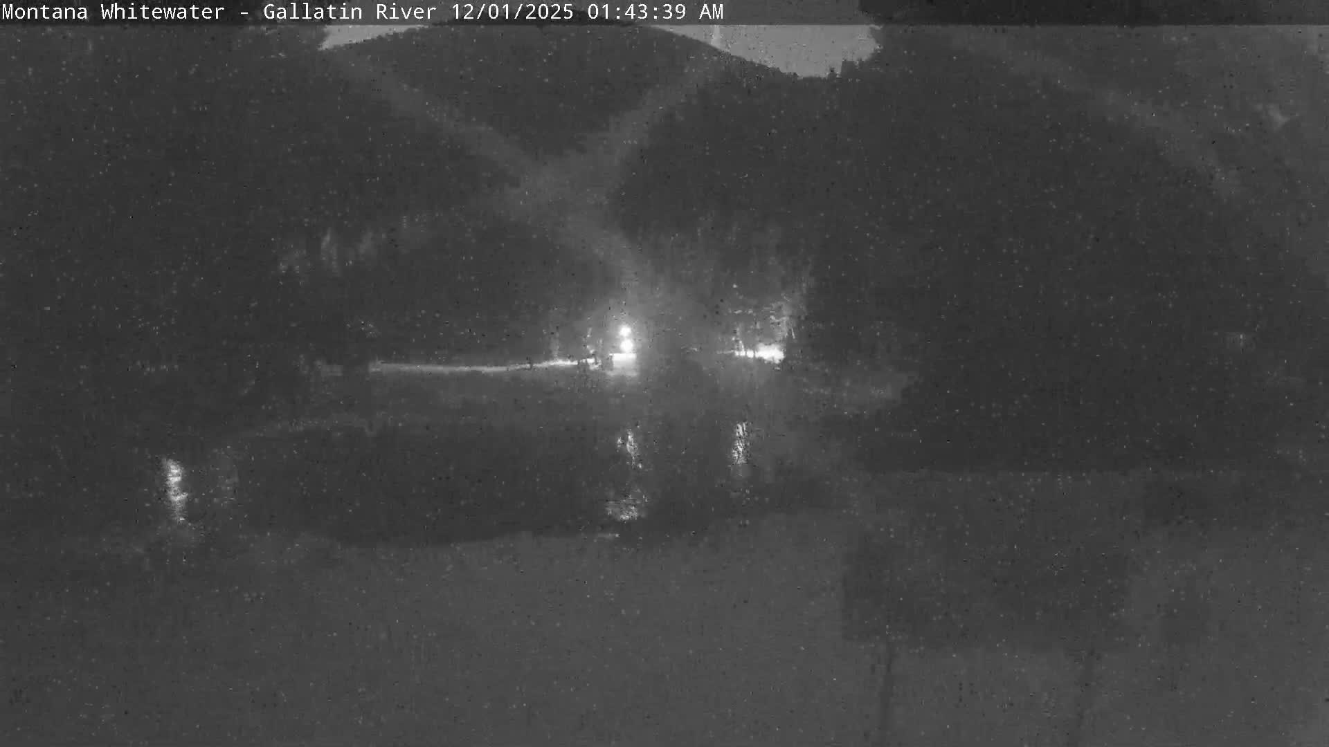 Bright lights reflect on a body of water at night against a dark, grainy background, with visible falling rain or snow.