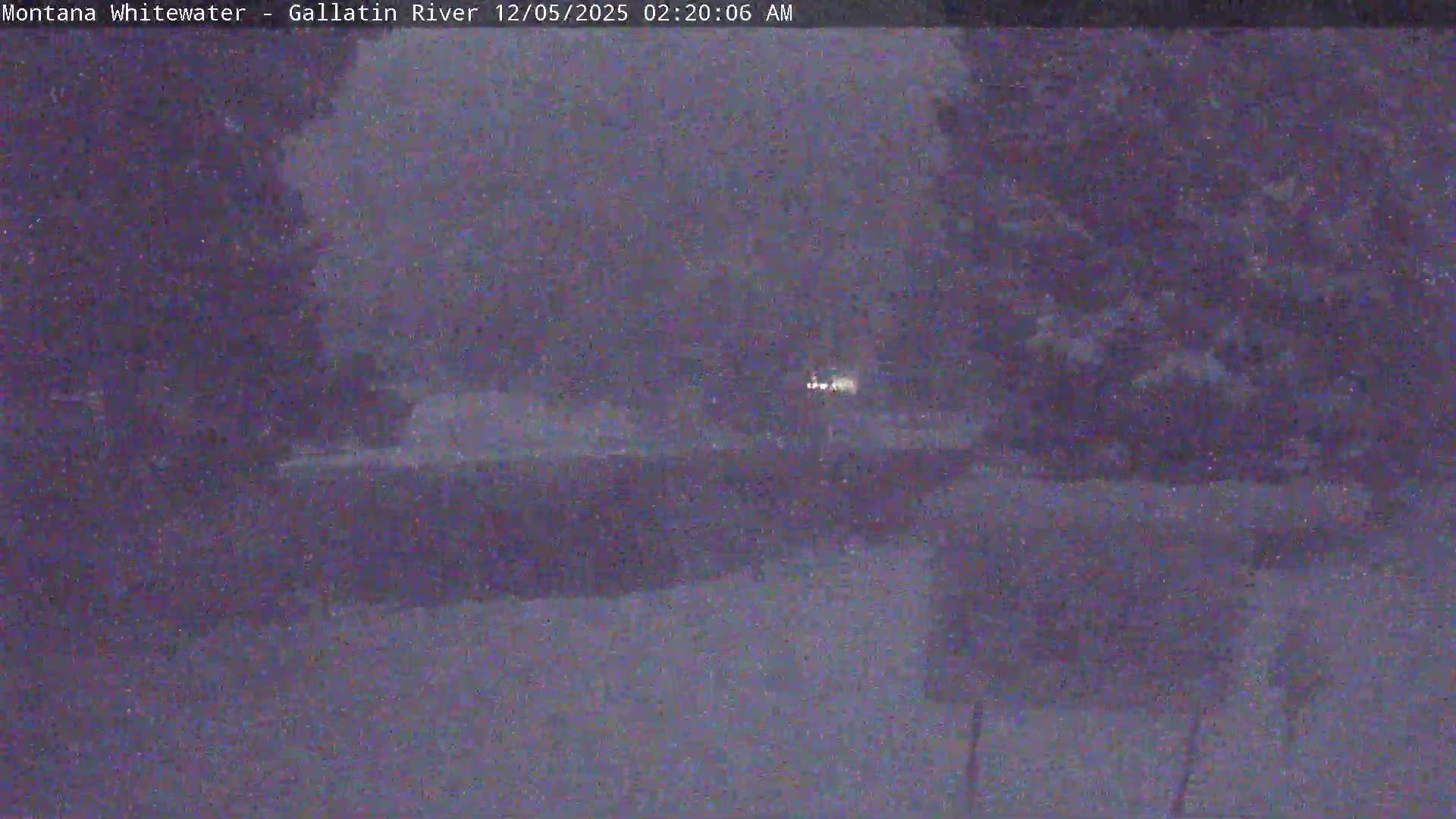 A dark, grainy nighttime image shows a wide river with faint distant lights on the far bank, partially obscured by what appears to be falling snow.