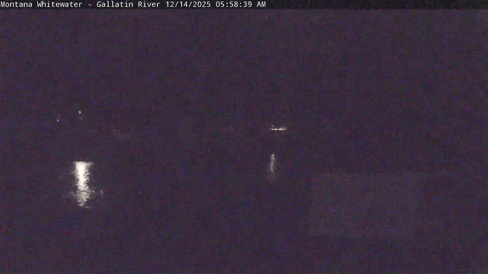 A dark, grainy nighttime image shows a wide river with faint distant lights on the far bank, partially obscured by what appears to be falling snow.