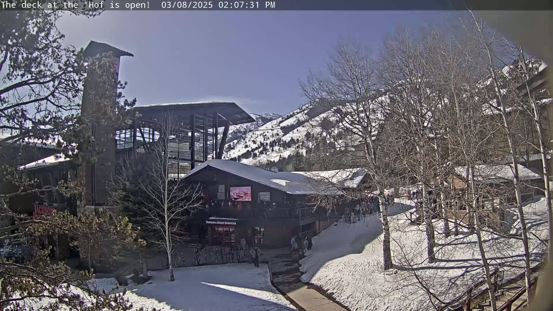A snow-covered outdoor scene on a sunny day shows a multi-story building with a deck, surrounded by bare trees and a snow-dusted mountain in the background.