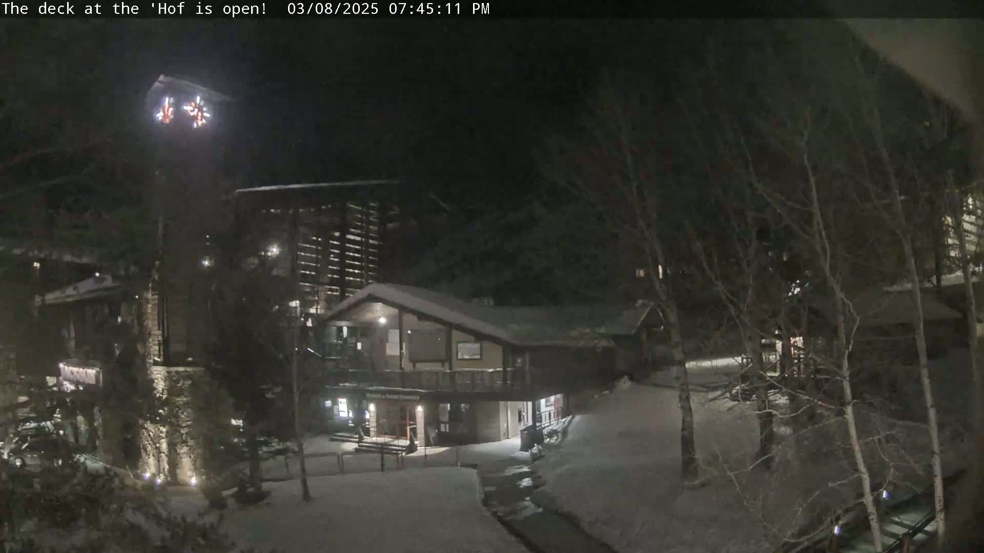 A nighttime snow-covered scene shows several buildings illuminated by artificial lights, surrounded by leafless trees.
