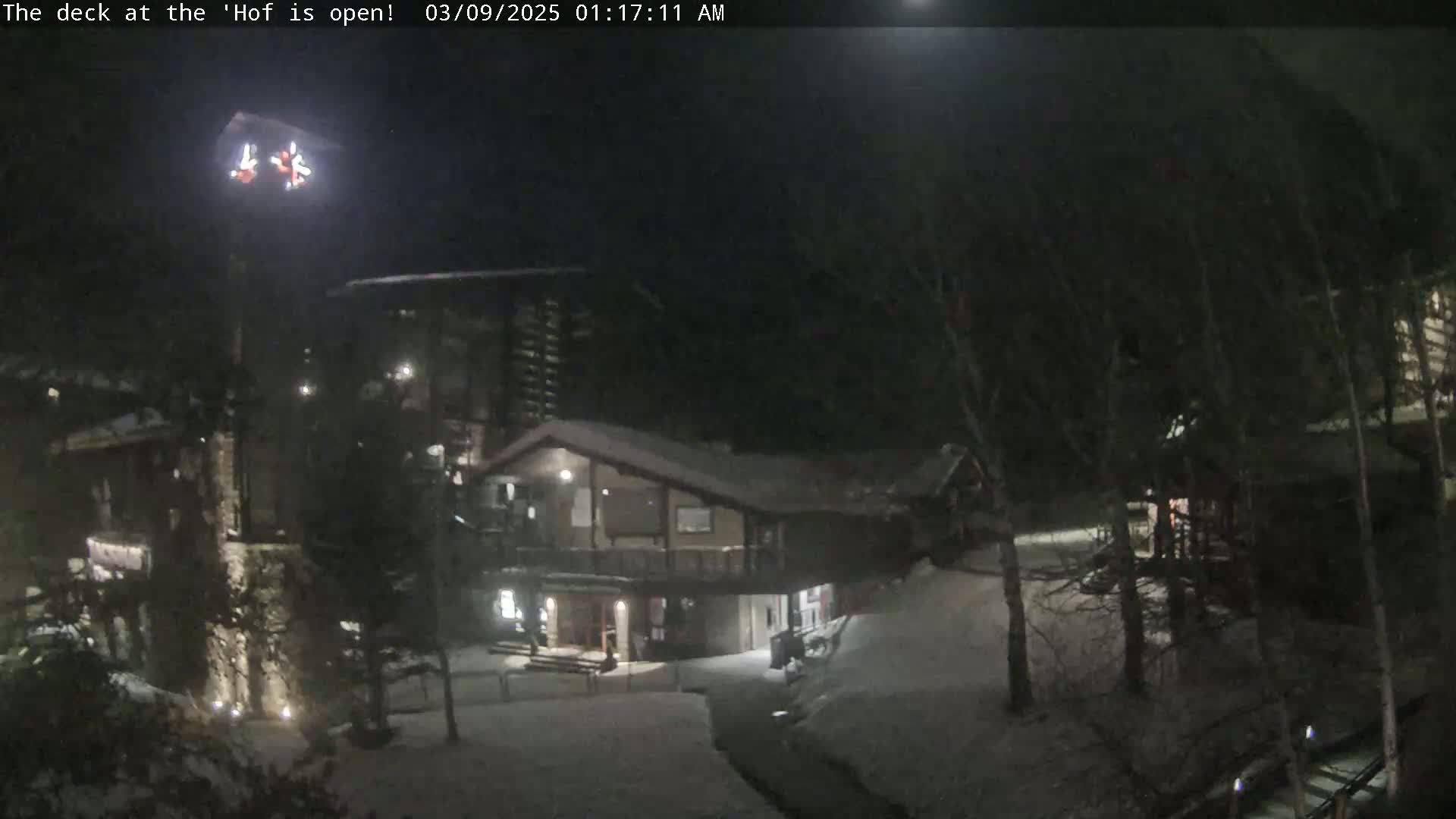 A nighttime snow-covered scene shows a multi-level building with exterior lighting, surrounded by trees.