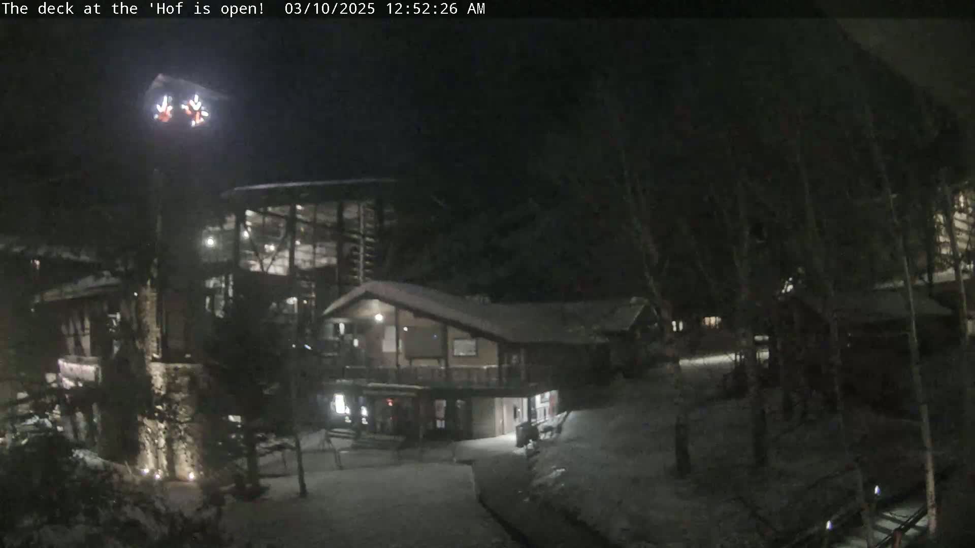 A snow-covered, nighttime scene shows a multi-level wooden building complex illuminated with artificial lights.