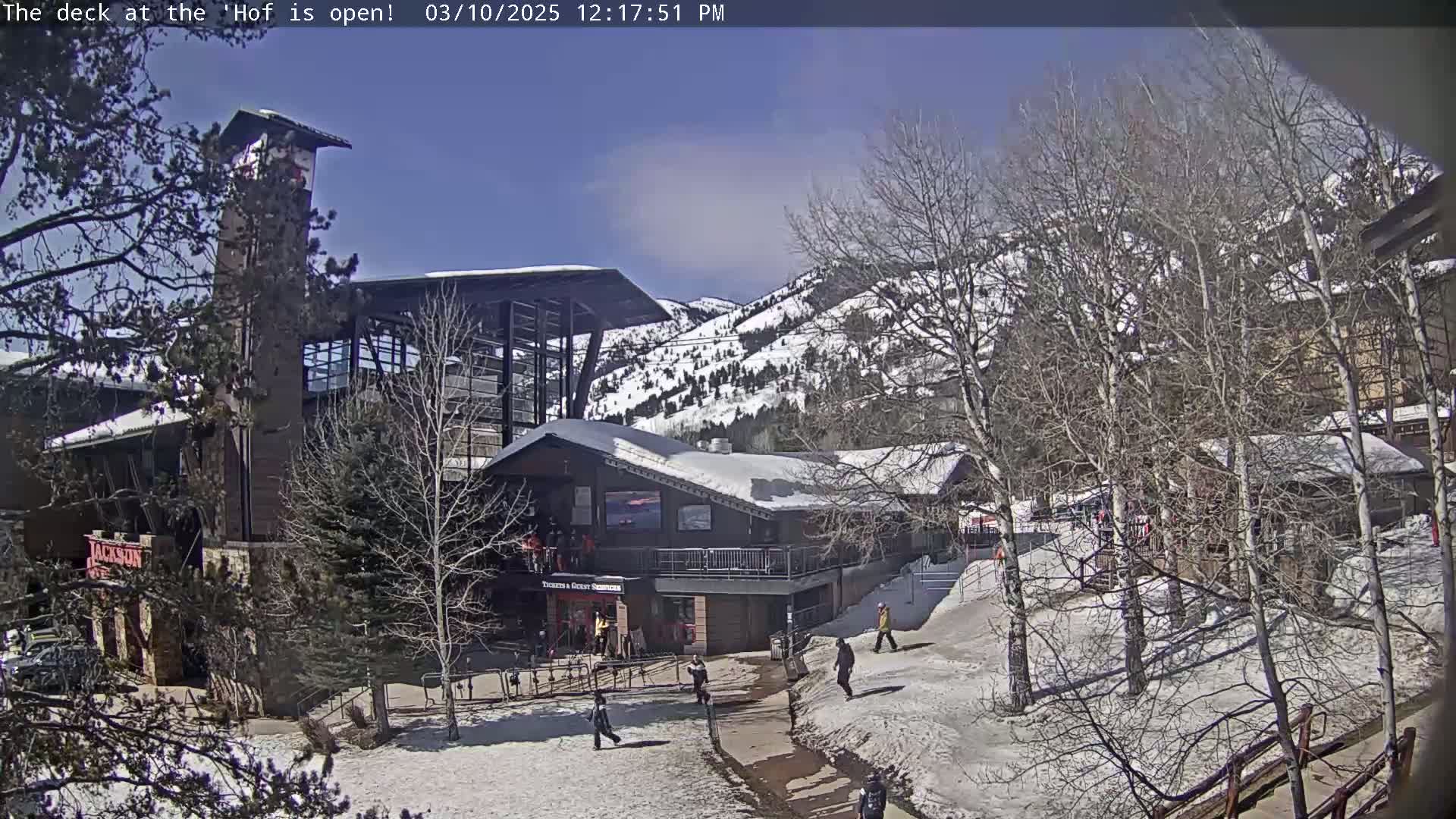 A snow-covered outdoor scene shows a cluster of buildings near bare trees and a snow-dusted mountainside under a mostly sunny sky.