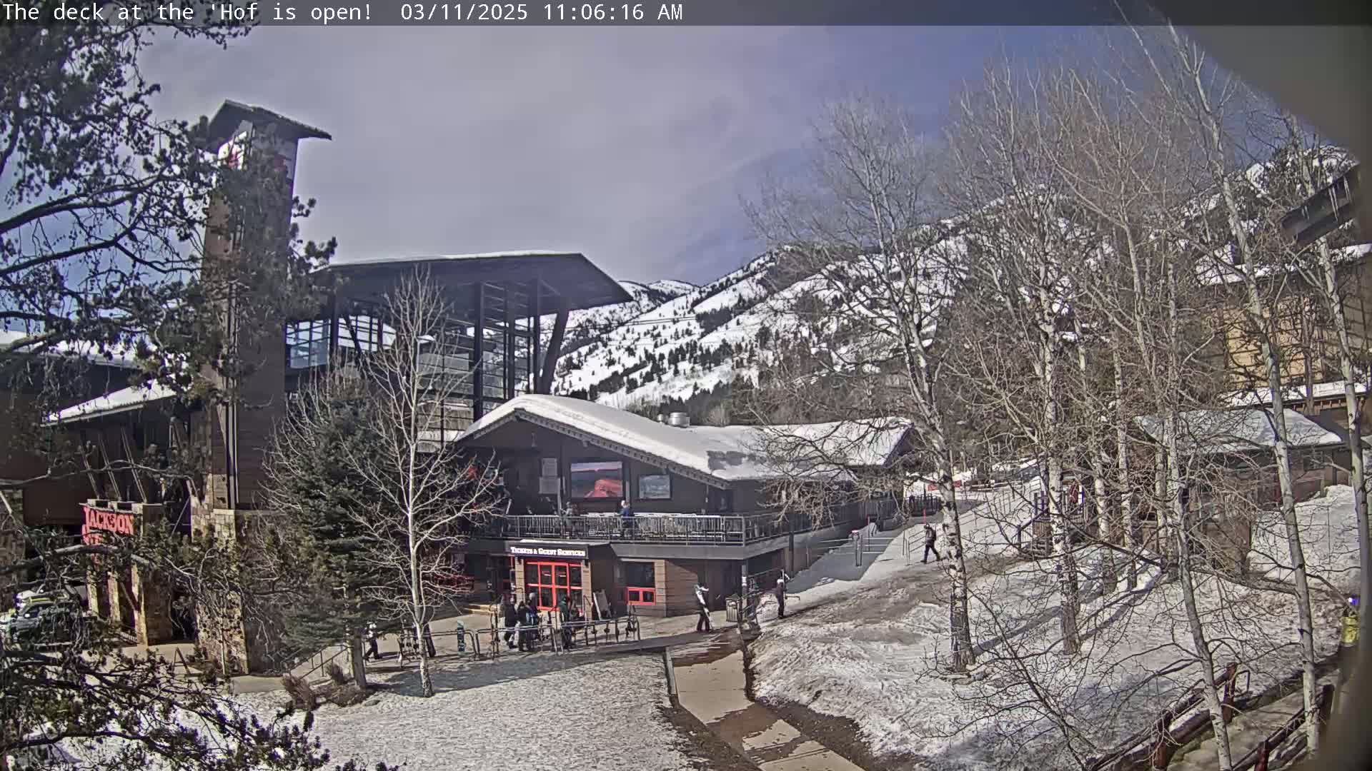 A snow-dusted mountainside with bare trees and several buildings, one with a large deck, under a mostly sunny sky.