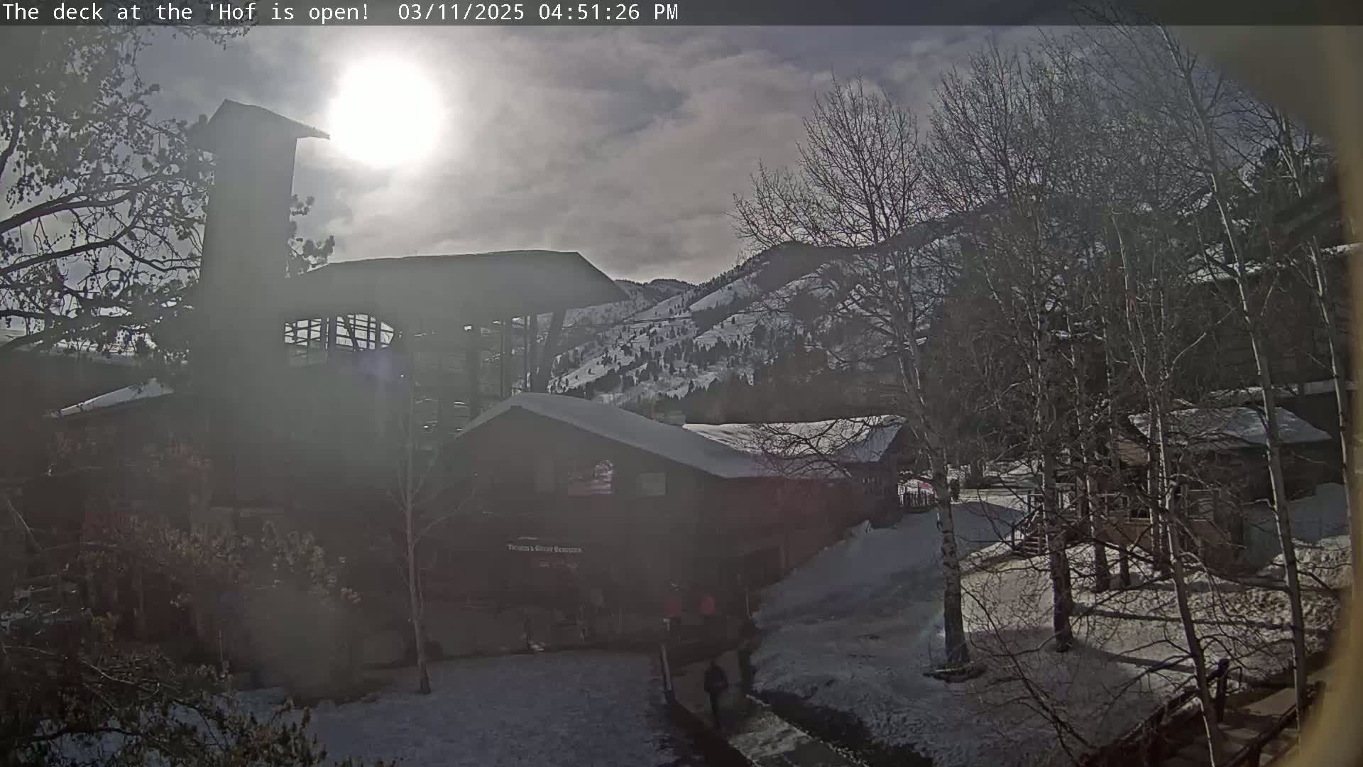 A sunny, partly cloudy day with snow on the ground shows a view of buildings and bare trees near a snow-covered mountainside.