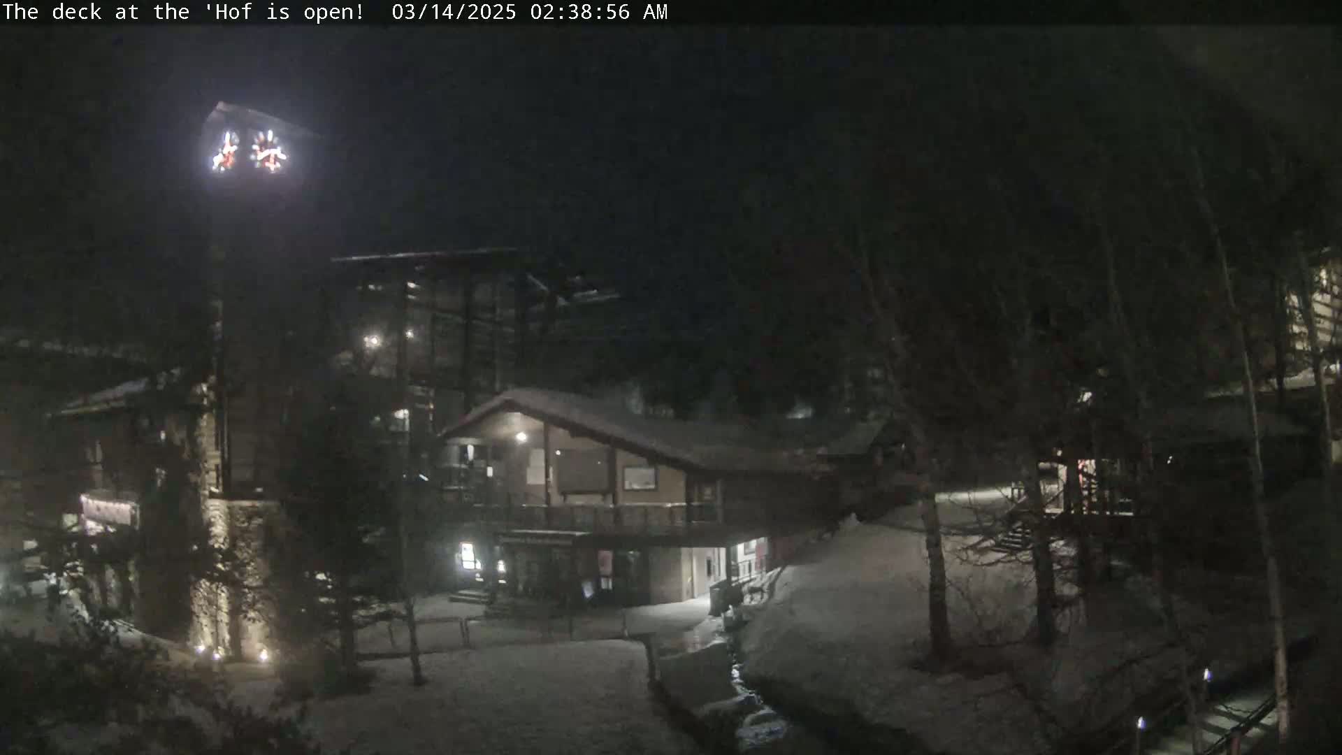 A nighttime view of a snow-covered area with buildings and trees, illuminated by artificial lights.