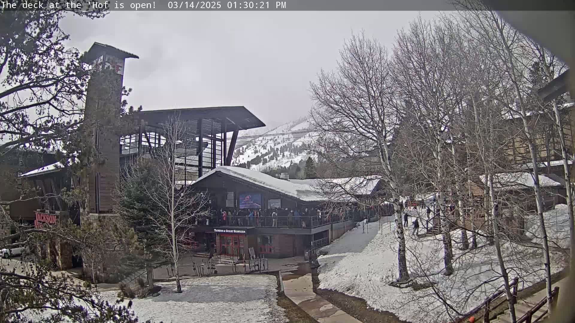 A snow-covered outdoor scene shows a multi-level building with people gathered on a deck, situated amongst bare trees and a snow-dusted mountain in the background under an overcast sky.