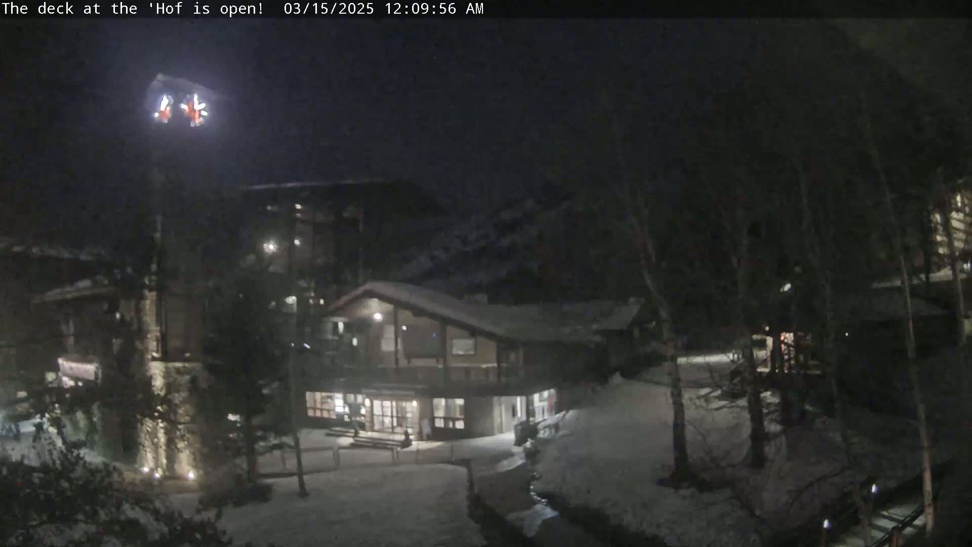 A snow-covered nighttime scene shows a lodge-like building with exterior lights illuminated, surrounded by trees and a slightly visible hill.