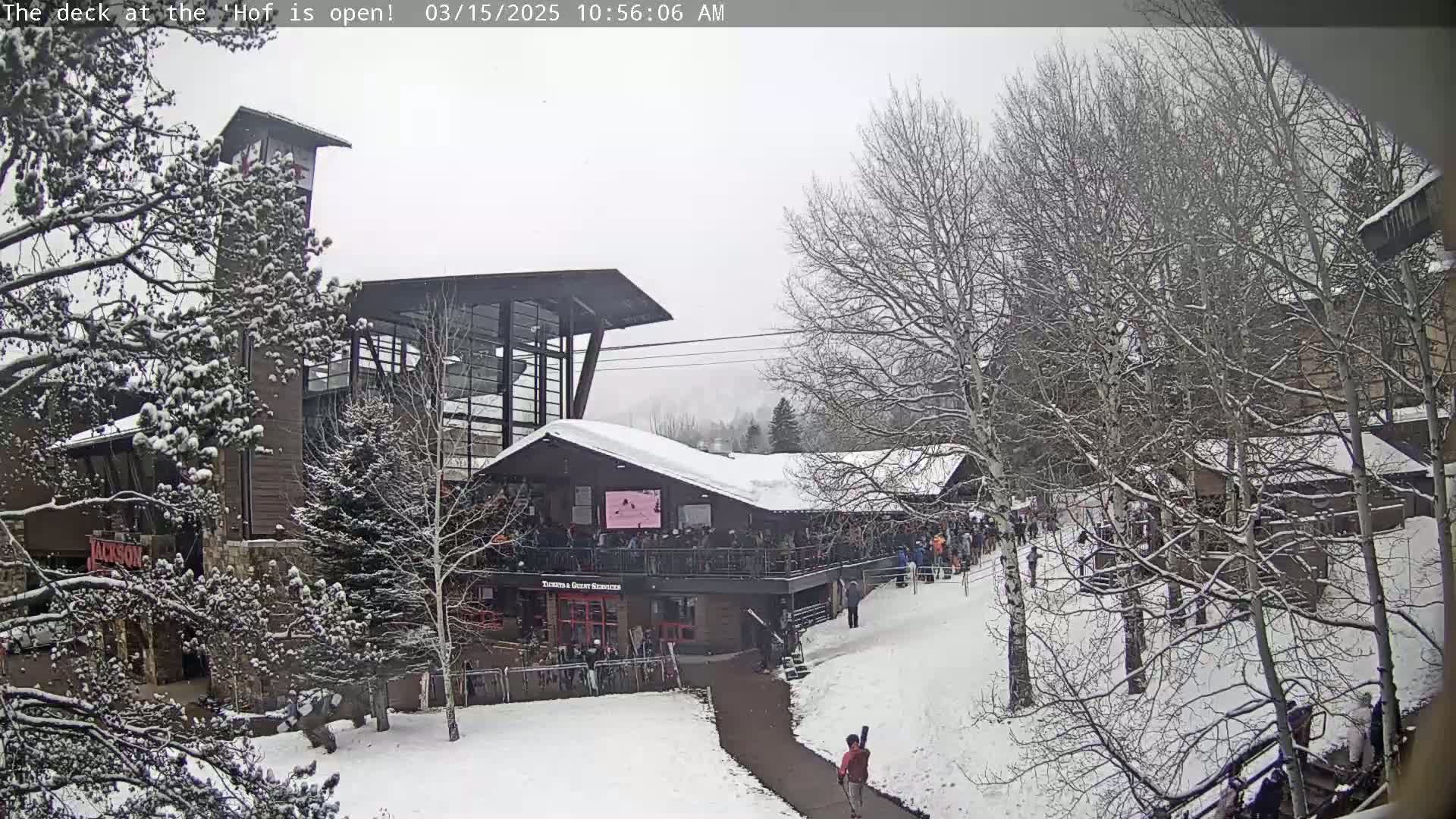 A snow-covered outdoor scene shows a multi-story building with a deck, surrounded by bare, snow-laden trees and a walkway with a few people.