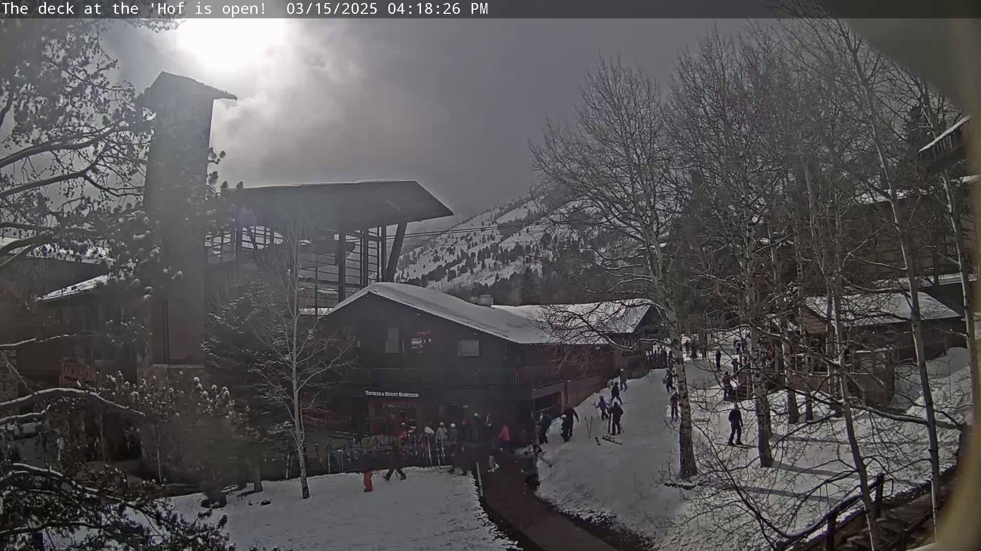 A snow-covered mountainside scene features several buildings, numerous people gathered near a structure, and leafless trees under a hazy, partly sunny sky.