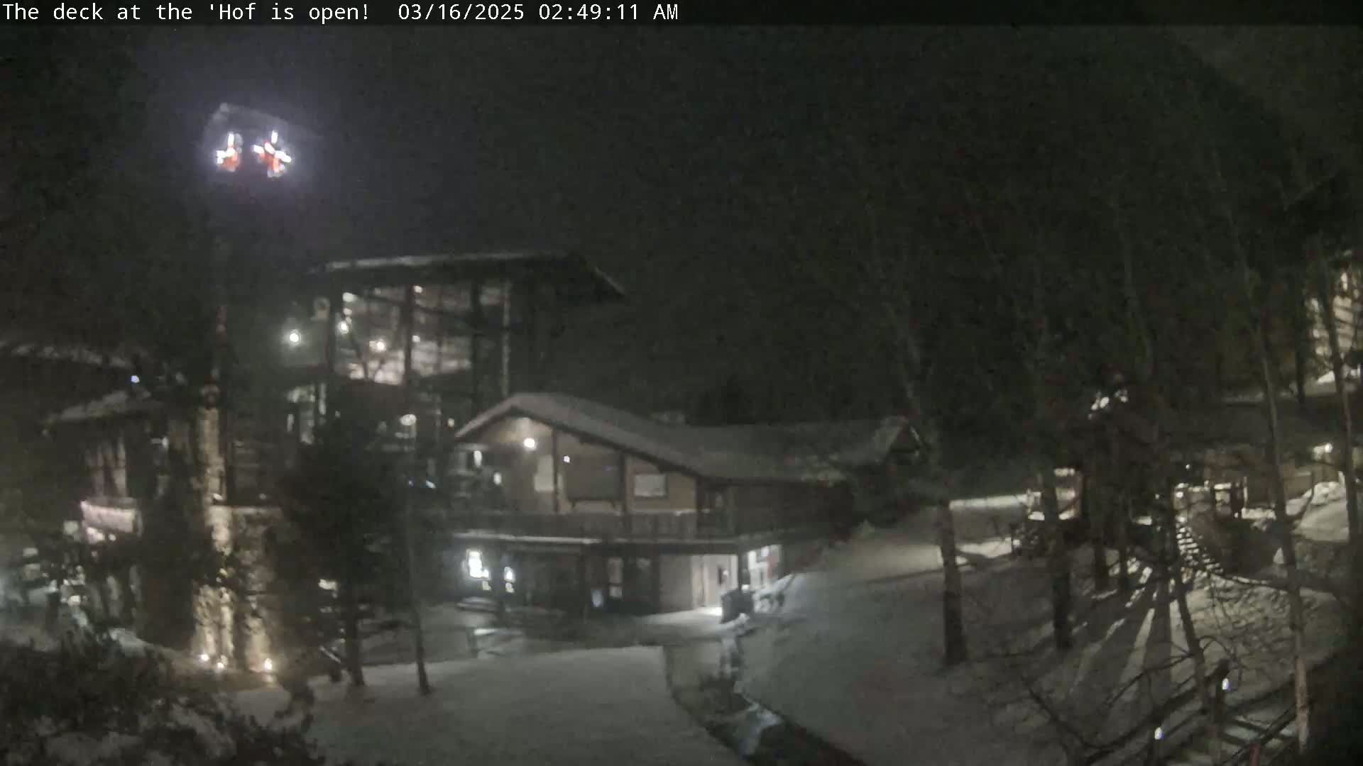 A nighttime snow scene shows two buildings illuminated, with snow covering the ground and trees.