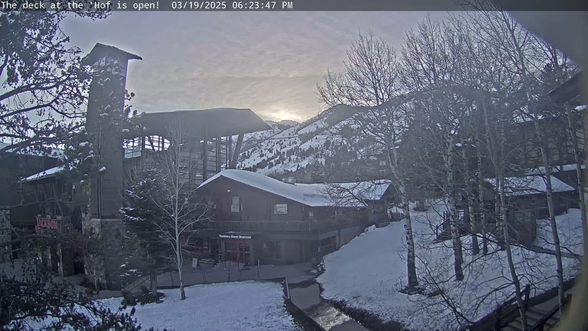 A snow-covered mountainside scene at dusk, featuring a complex of buildings with a tall tower, and bare trees in the foreground.