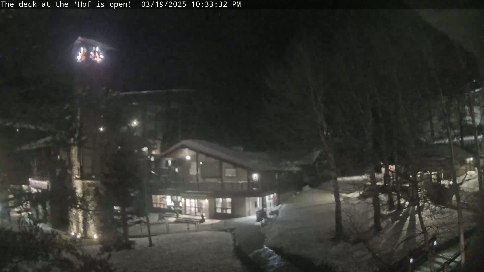 A snow-covered nighttime scene shows a building with a deck, illuminated by lights, surrounded by trees.