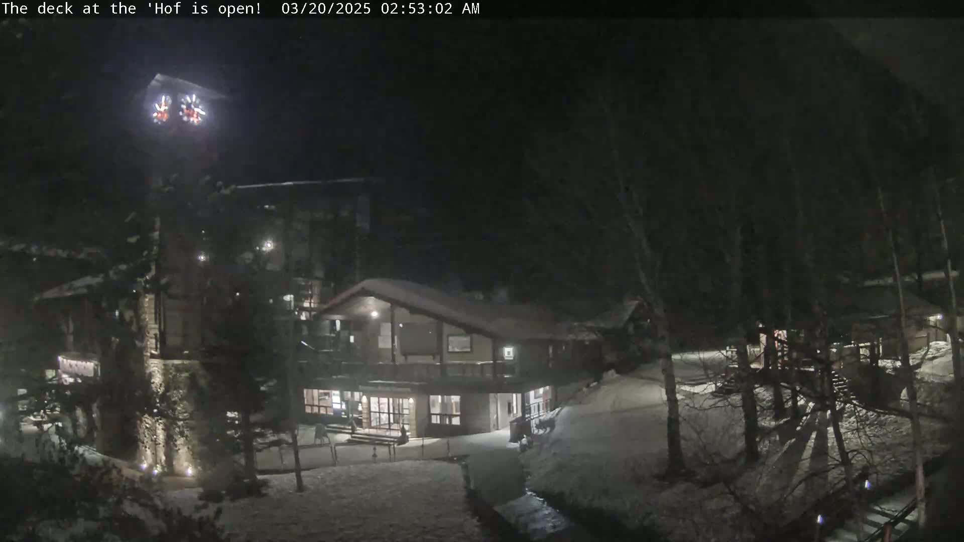A snow-covered nighttime scene shows a multi-level building with exterior lighting, surrounded by trees.