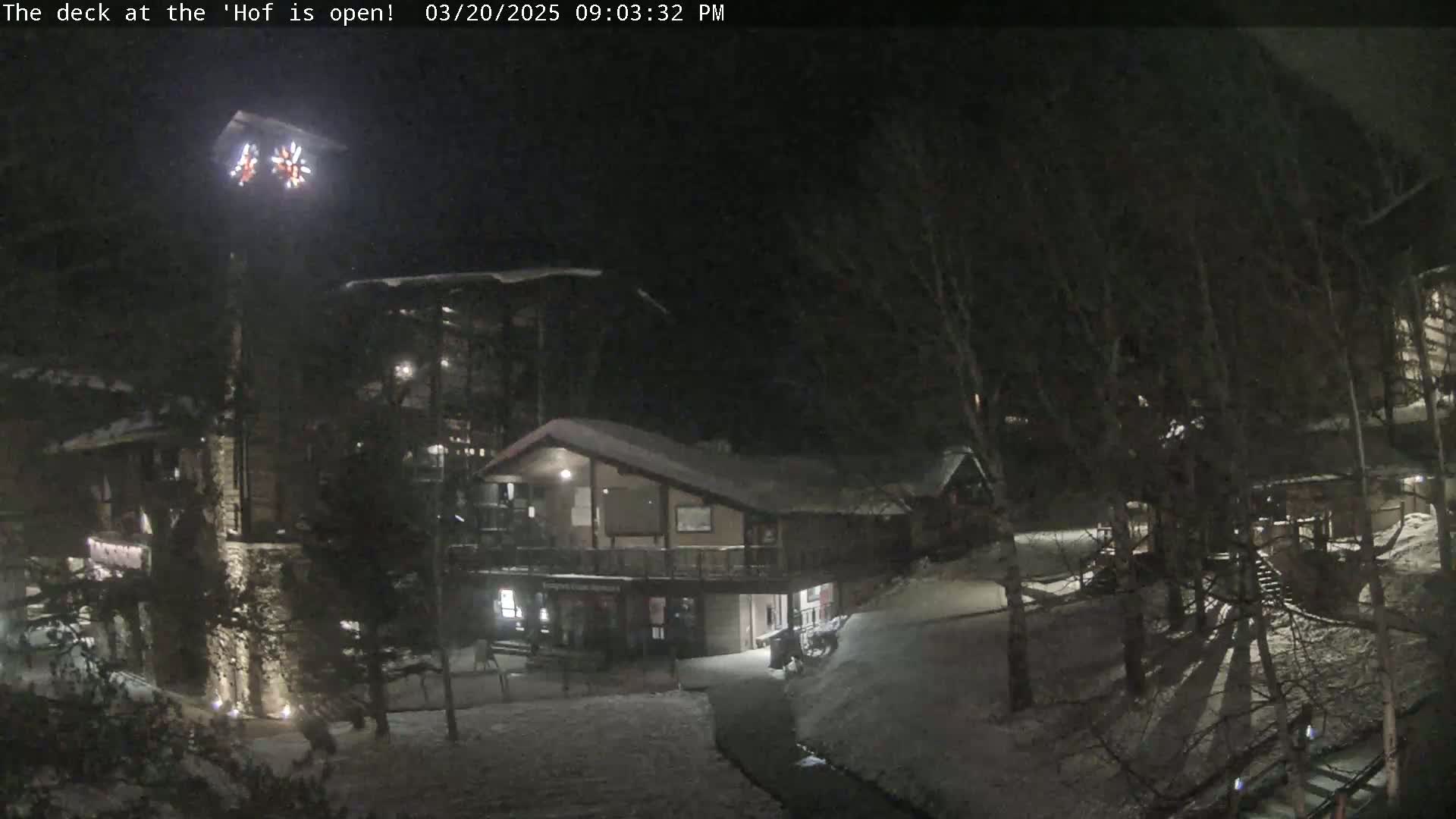 A snow-covered outdoor area at night is illuminated, showing a multi-story building with a smaller structure connected to it, surrounded by trees.