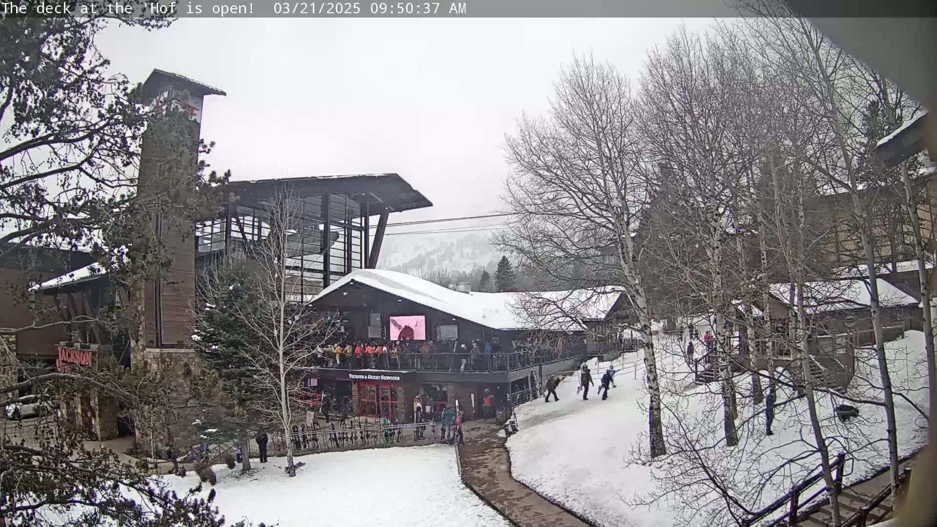 A snow-covered outdoor scene shows a multi-story building with a deck and people, under a gray, overcast sky.