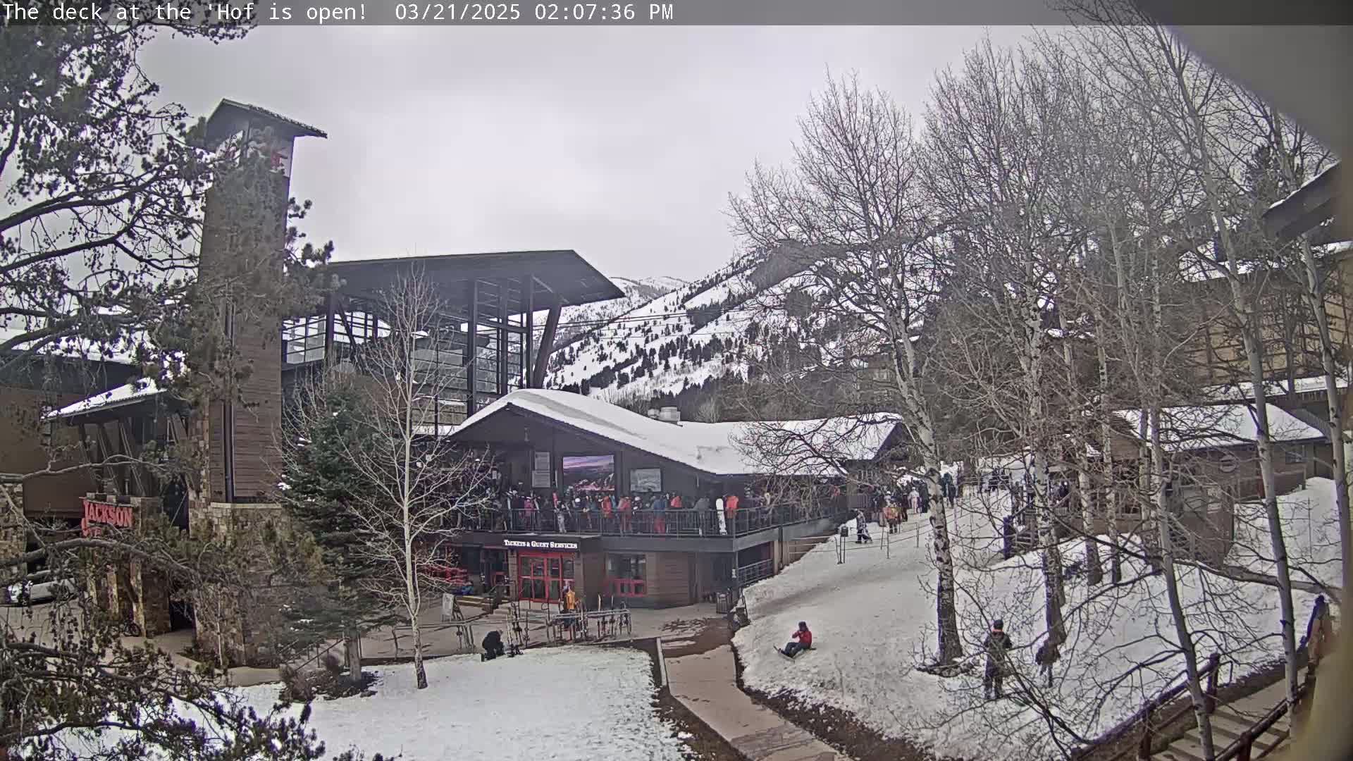 A snow-covered outdoor area with a multi-story building, a crowd of people on a deck, and bare trees under an overcast sky.