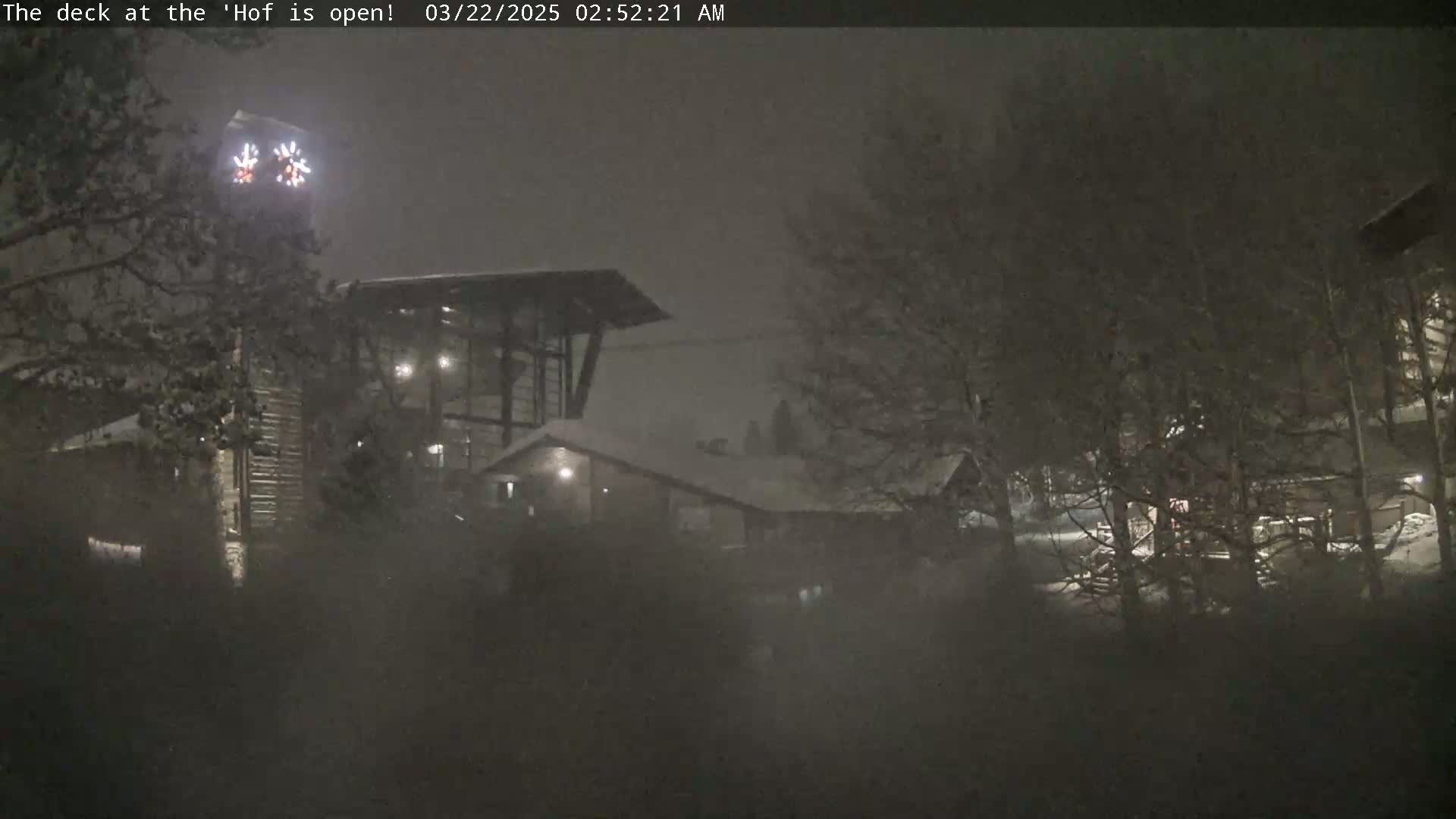 Snow falls at night on a snow-covered building complex partially obscured by trees.