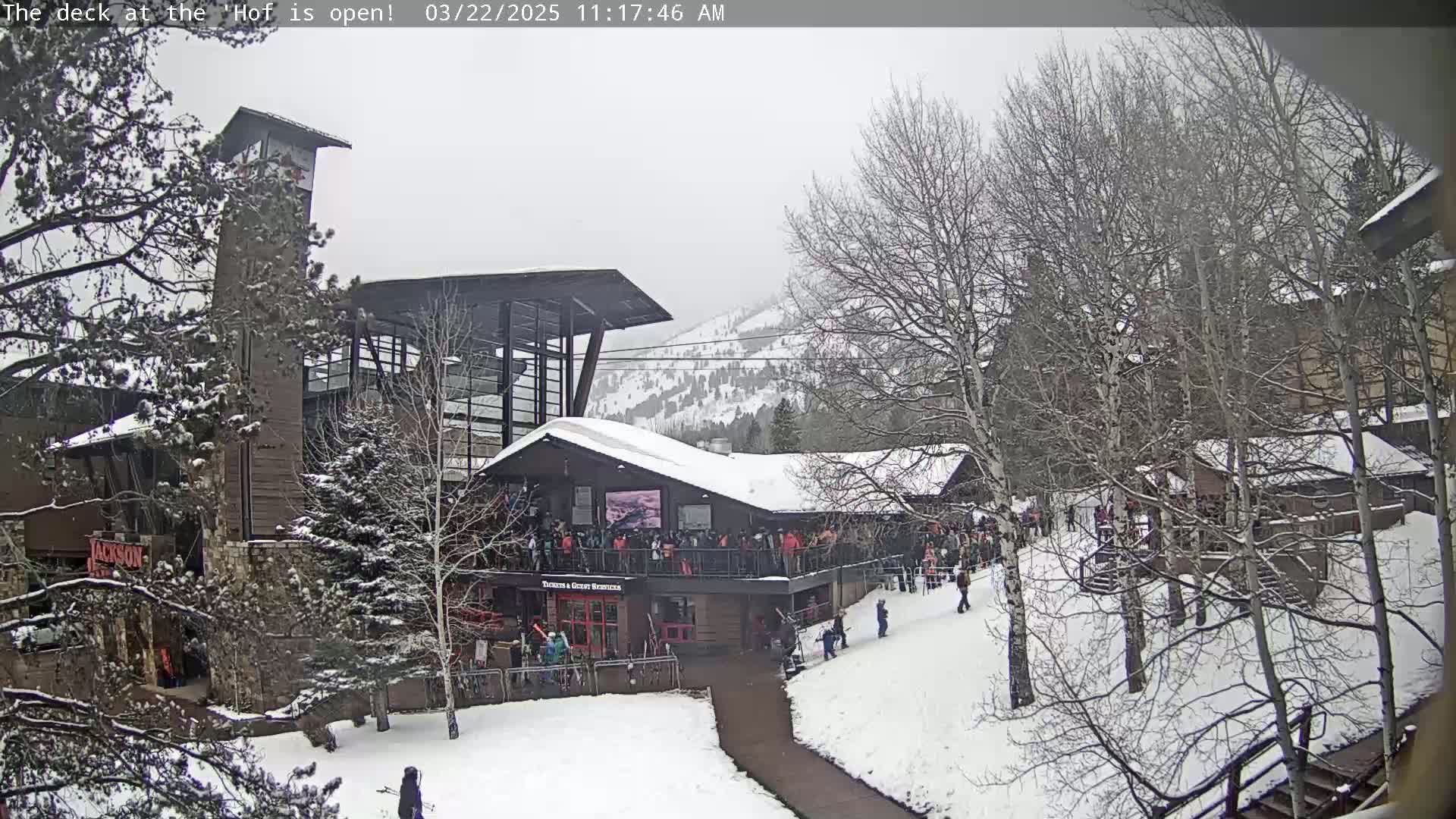 A snow-covered outdoor scene shows a multi-story building with a deck crowded with people, surrounded by bare trees and a snow-covered landscape under an overcast sky.
