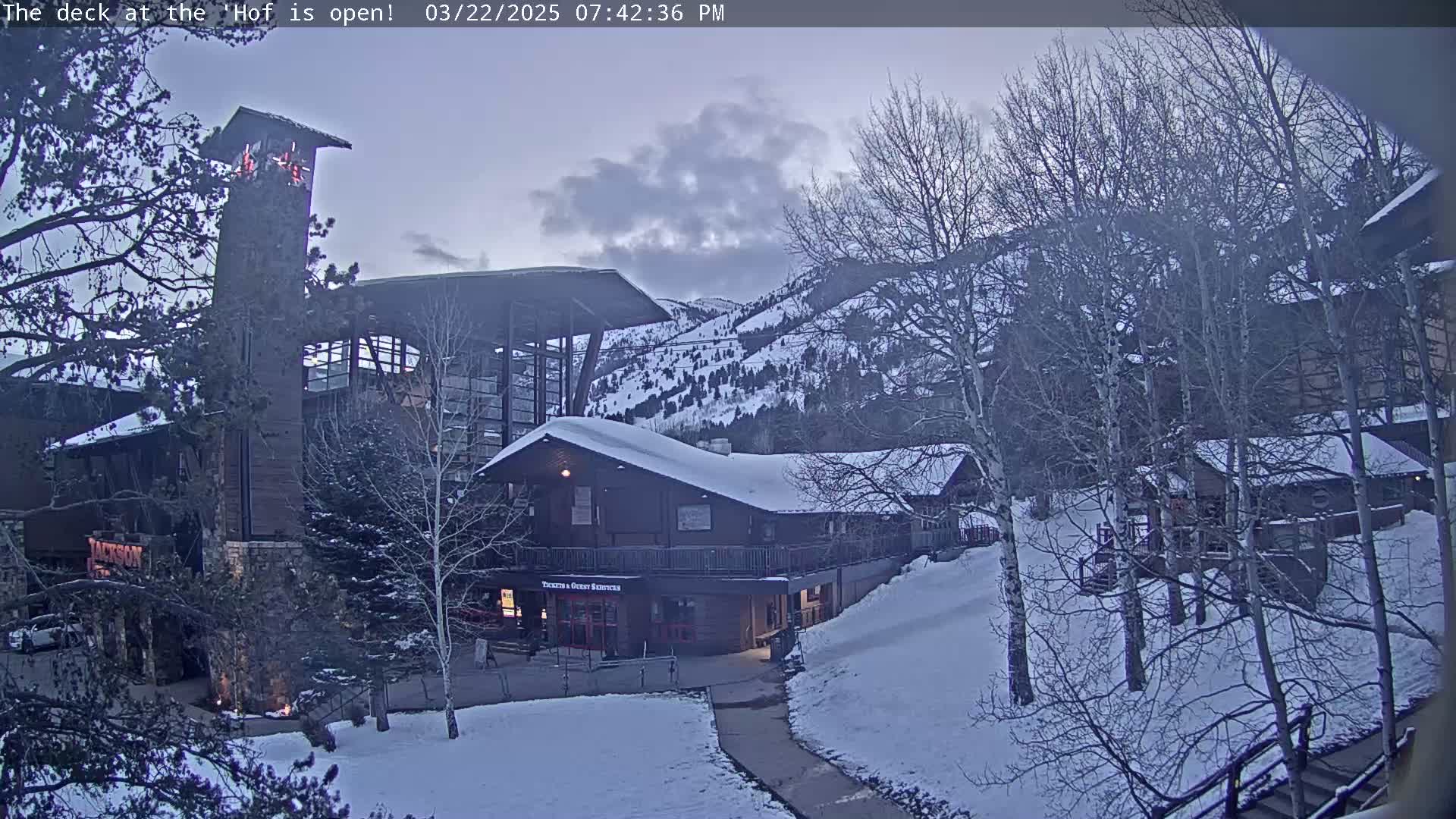 A snow-covered resort complex sits beneath a partly cloudy twilight sky, with bare trees surrounding the buildings and a mountain range in the background.
