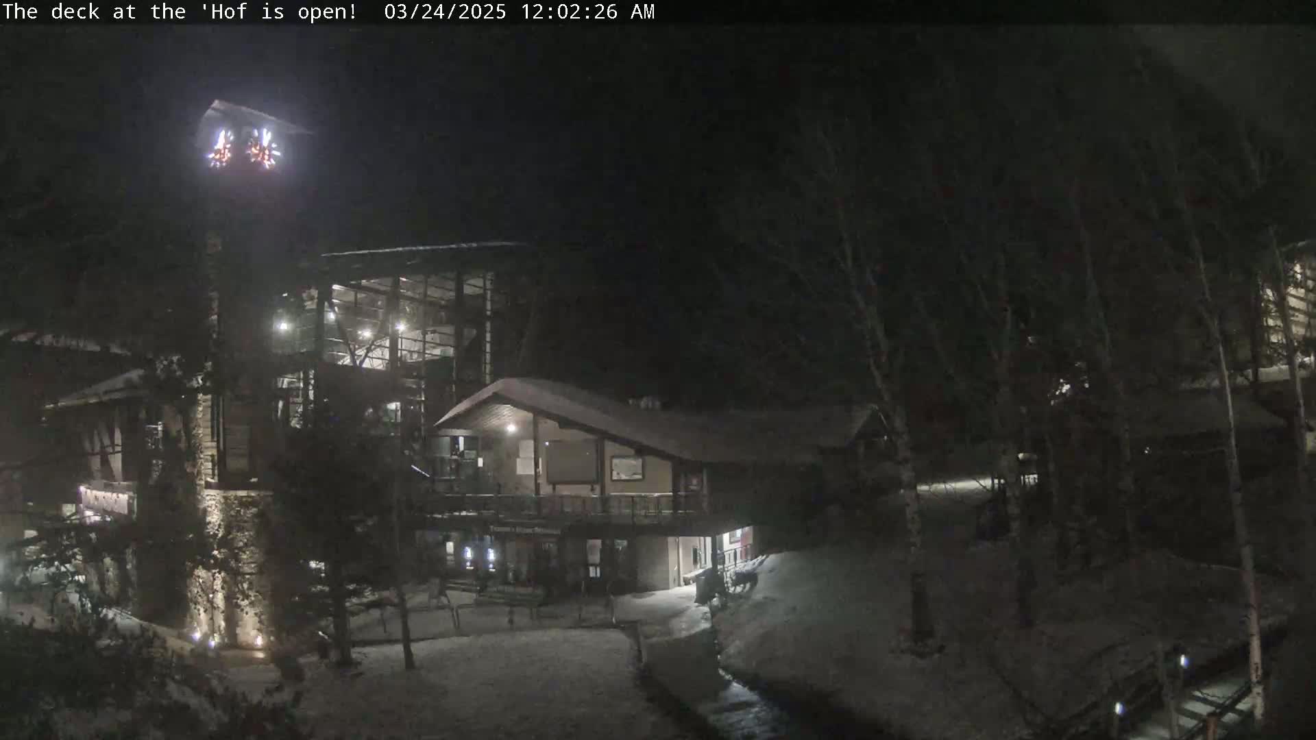 A nighttime snow-covered scene shows a multi-level building with lights on, partially obscured by trees and a small stream.