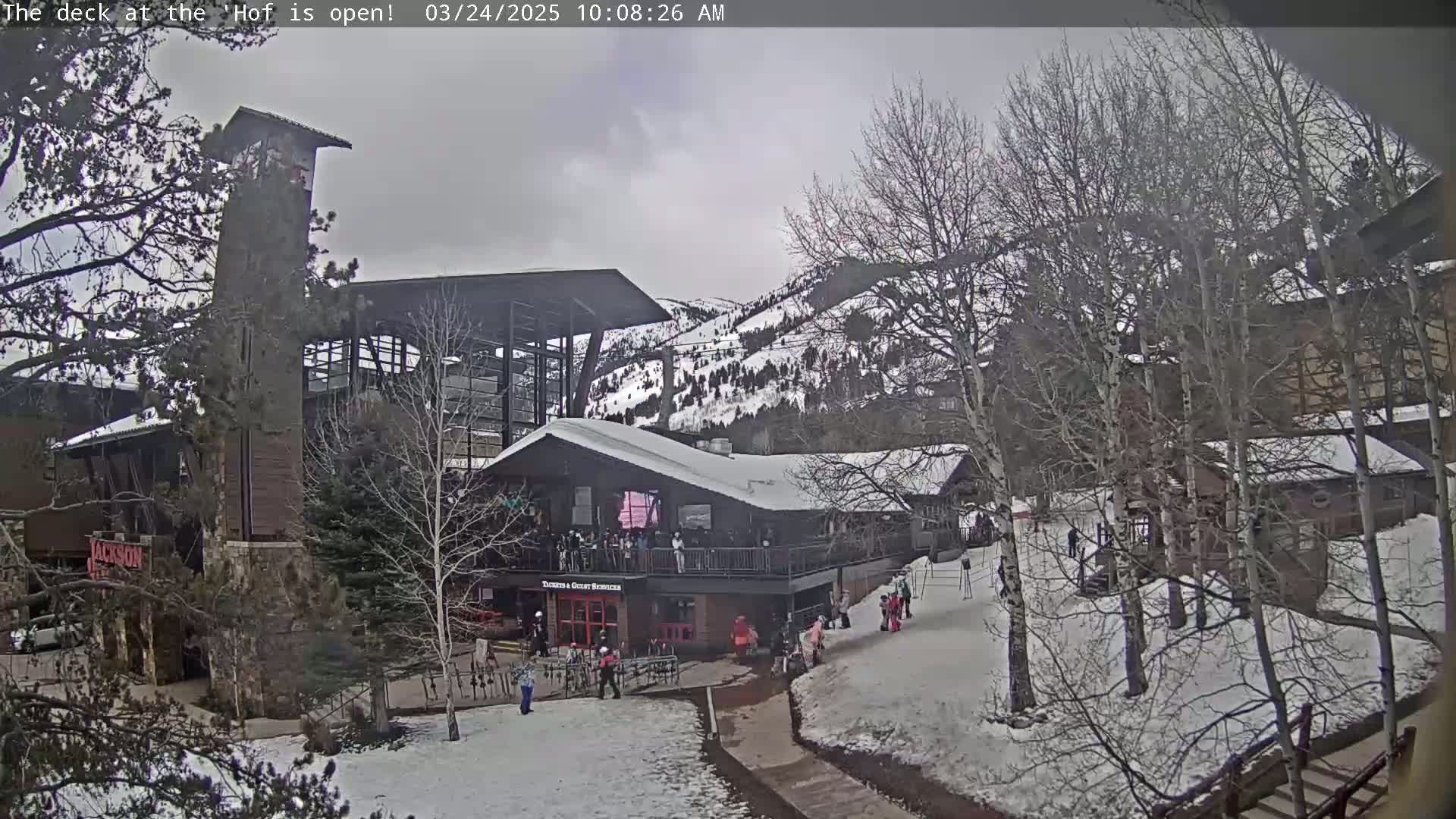 A snow-covered outdoor scene shows a multi-story building complex nestled amongst bare trees and a snow-dusted mountainside under a cloudy sky.