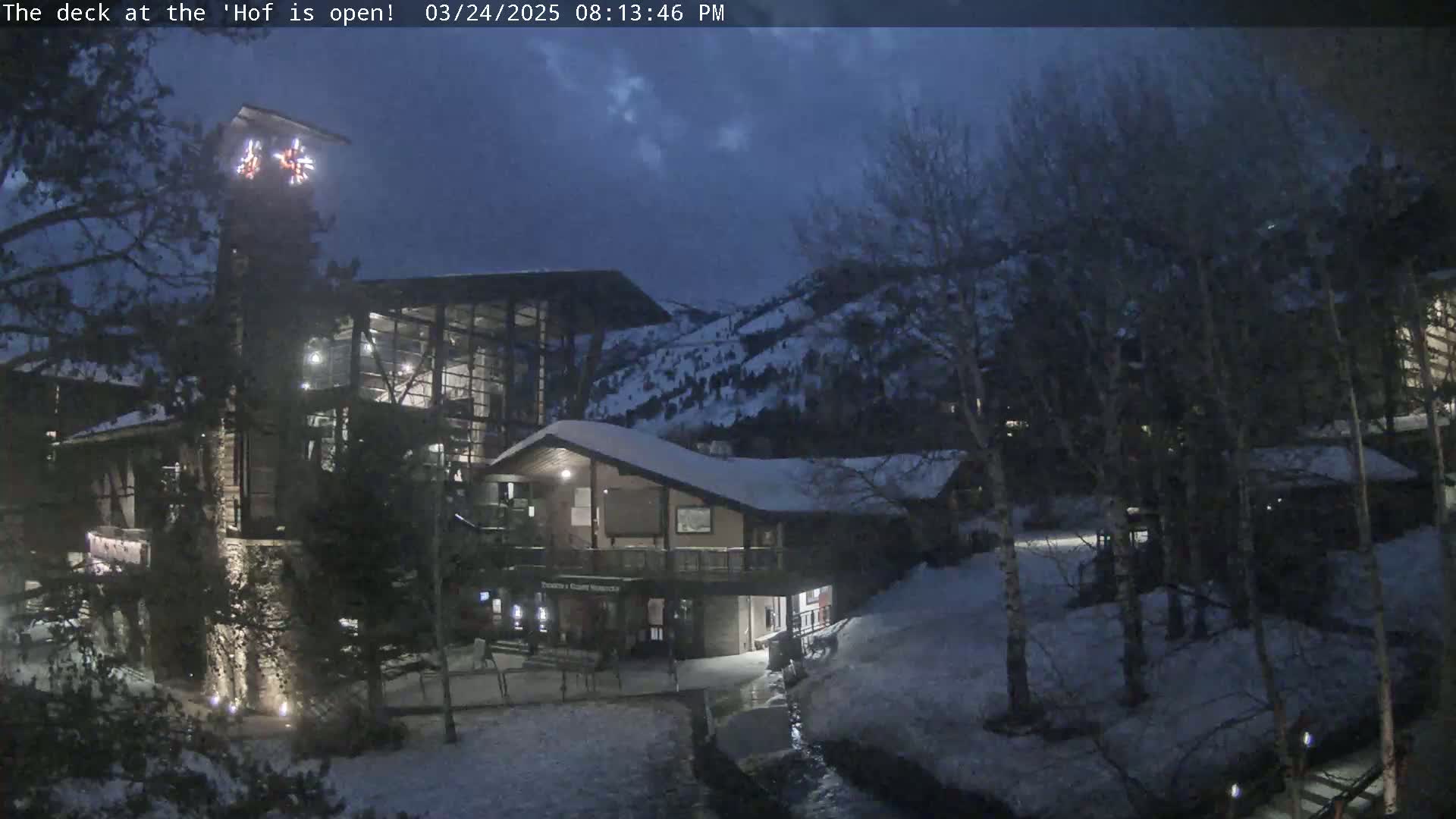A snow-covered complex of buildings with large windows is illuminated at night, under a dark twilight sky, with a mountainside visible in the background.