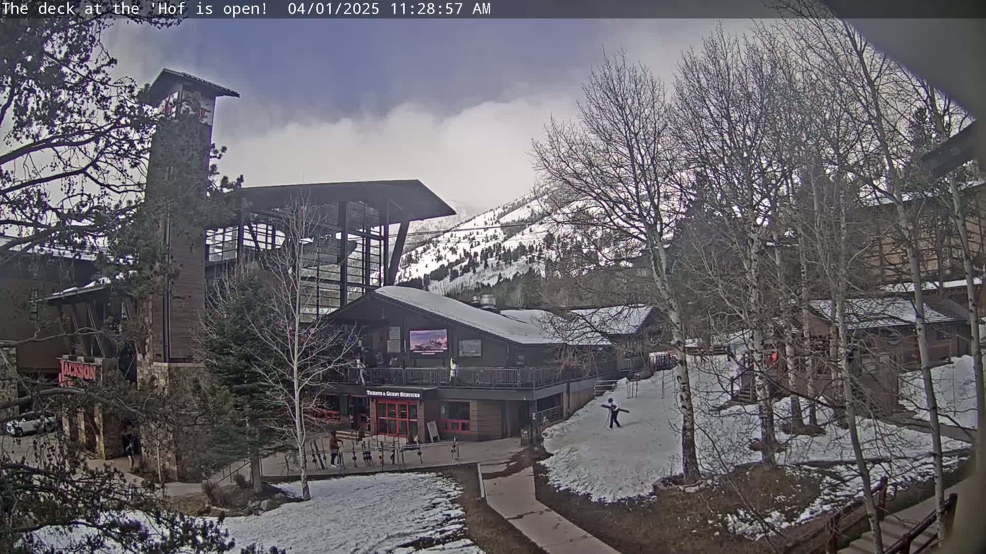 A snow-covered mountainside is visible behind a multi-story building complex with a deck, under an overcast sky.
