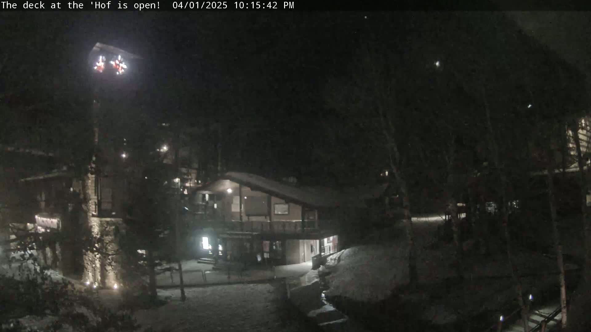 A nighttime view of a snow-covered lodge-like building with some lights on, partially surrounded by trees under a dark, possibly snowy sky.