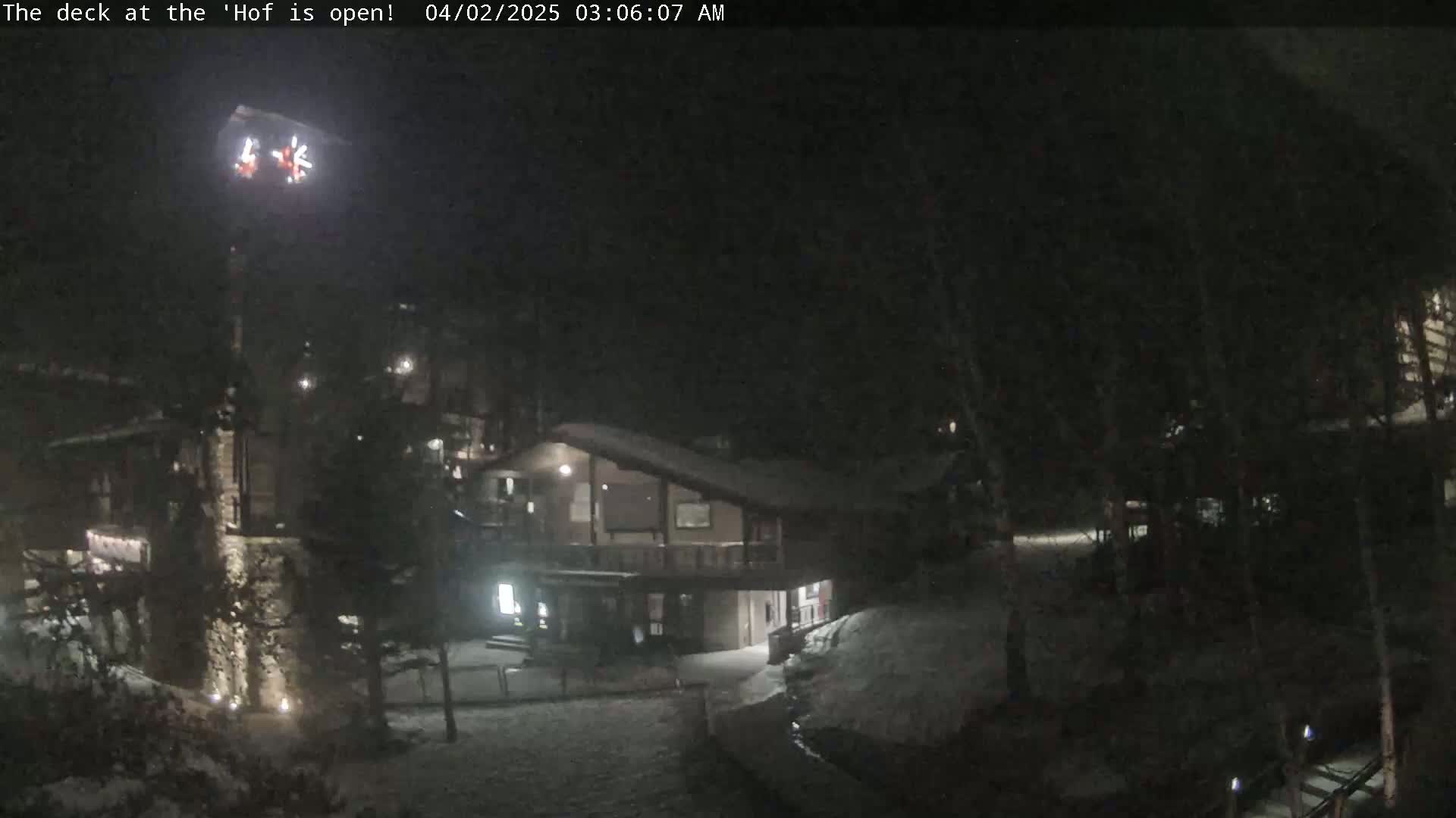 A snow-covered nighttime scene shows a two-story building with outdoor lights partially obscured by trees.