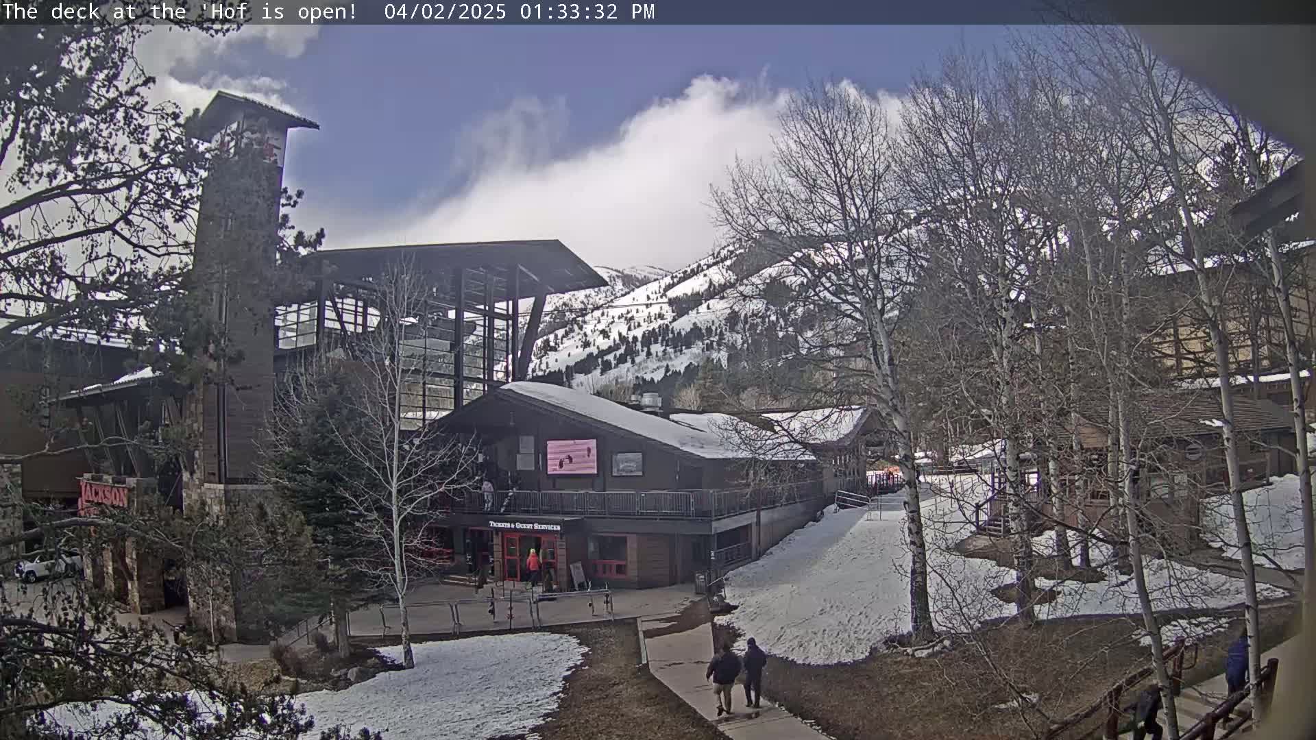 A partly cloudy day reveals a mountainside resort with snow-covered ground and leafless trees surrounding buildings.