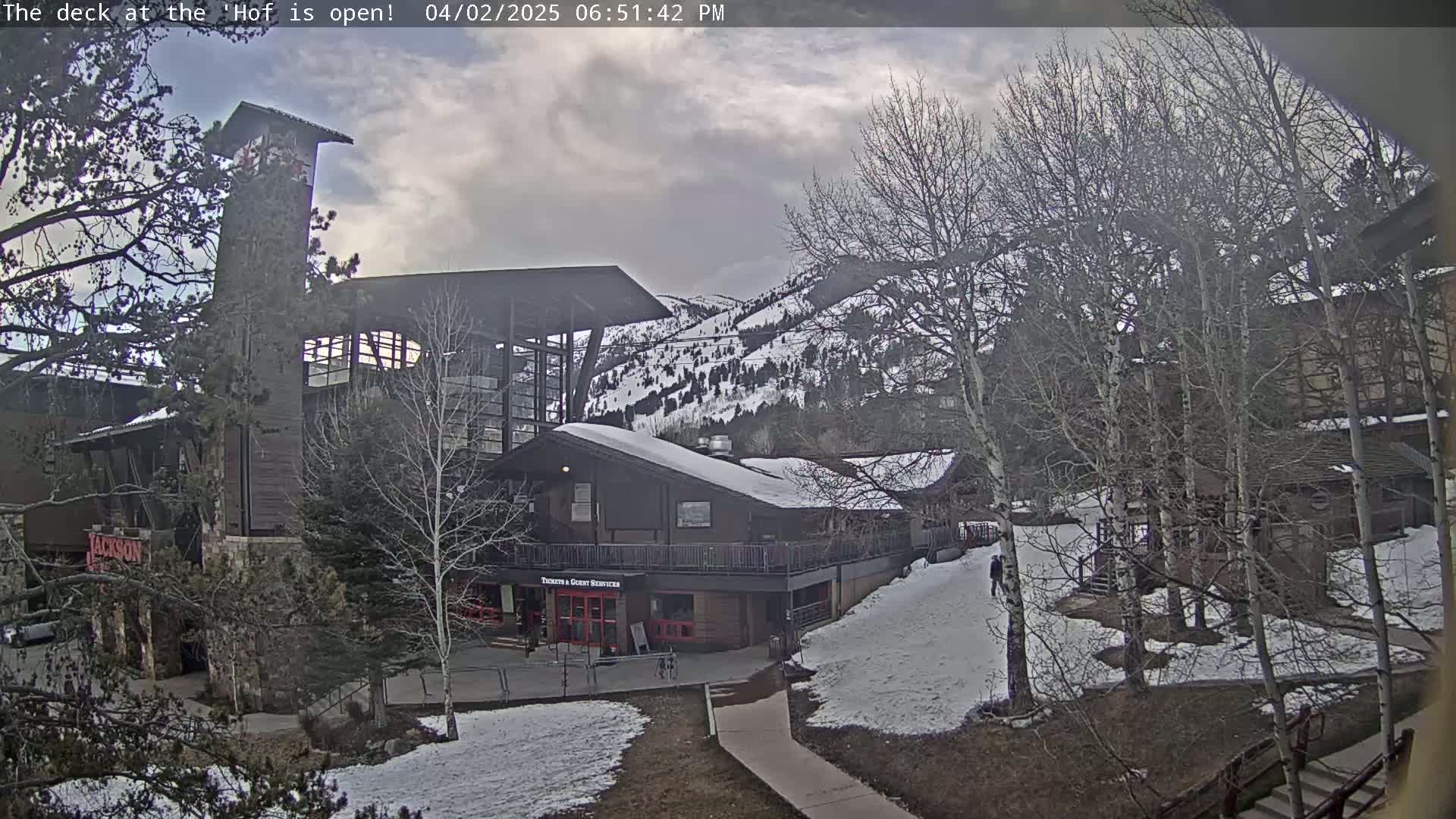 A partially snow-covered mountainside with a building complex under a cloudy sky.