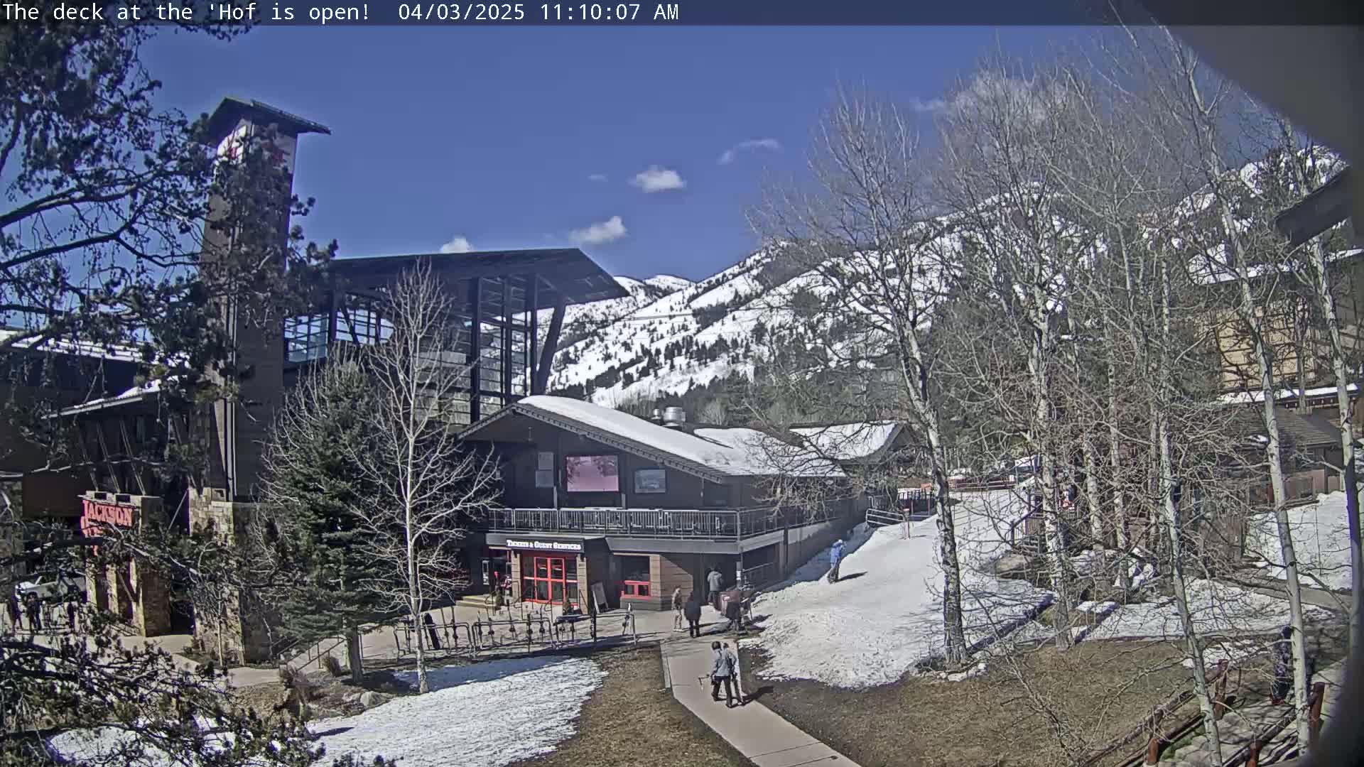 A sunny day reveals a snow-dusted landscape with a multi-story building complex, surrounded by leafless trees and a mountain backdrop partially covered in snow.