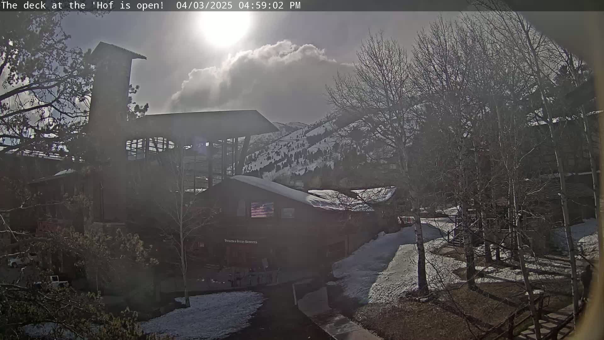 A partly sunny day with snow on the ground shows several buildings nestled among leafless trees and snow-covered hills in the background.