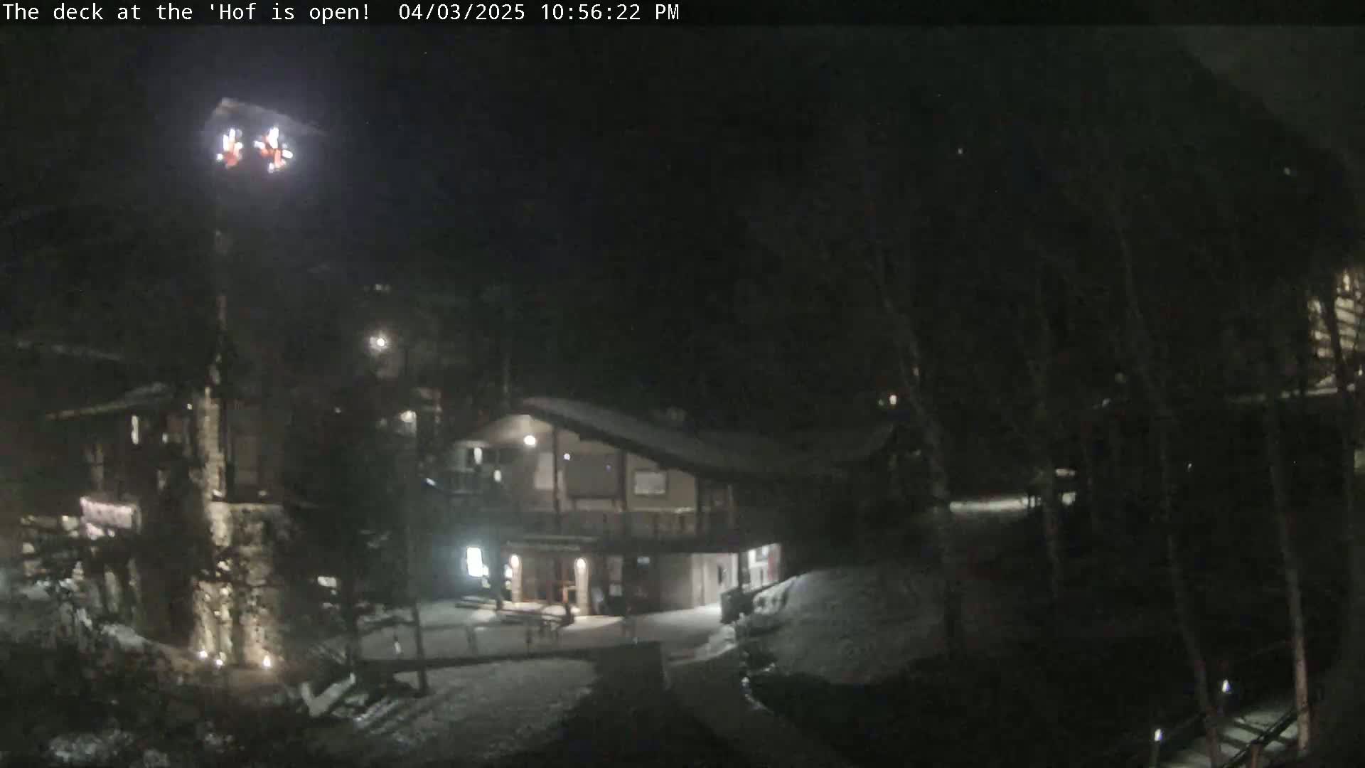 A nighttime, snow-covered scene shows a building with lights on, surrounded by dark trees under a dark sky.