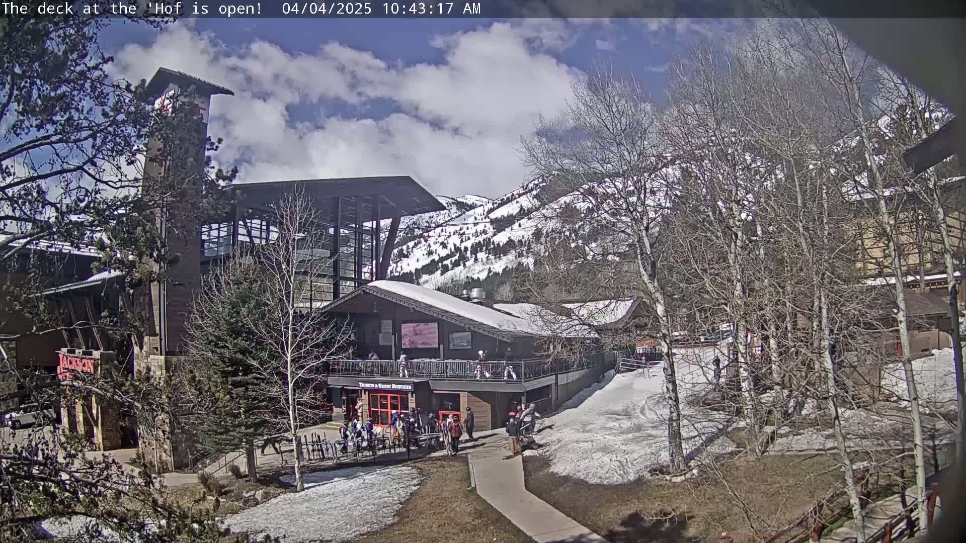 A partially snow-covered outdoor deck and building complex are situated against a backdrop of snow-dusted mountains under a partly sunny sky.