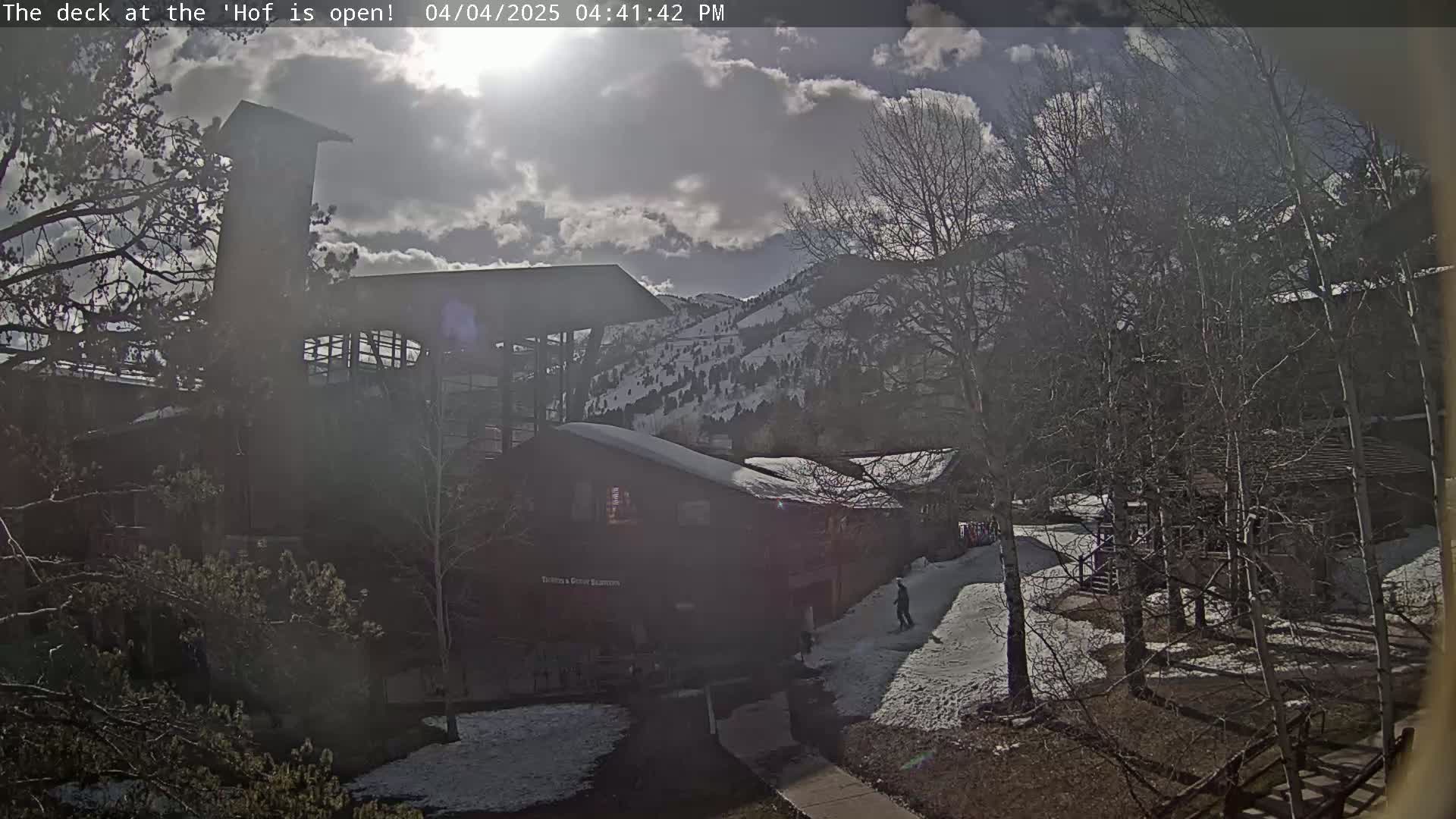 A partly sunny day with some snow on the ground shows a view of several buildings nestled among leafless trees and snow-dusted ground against a backdrop of mountains.